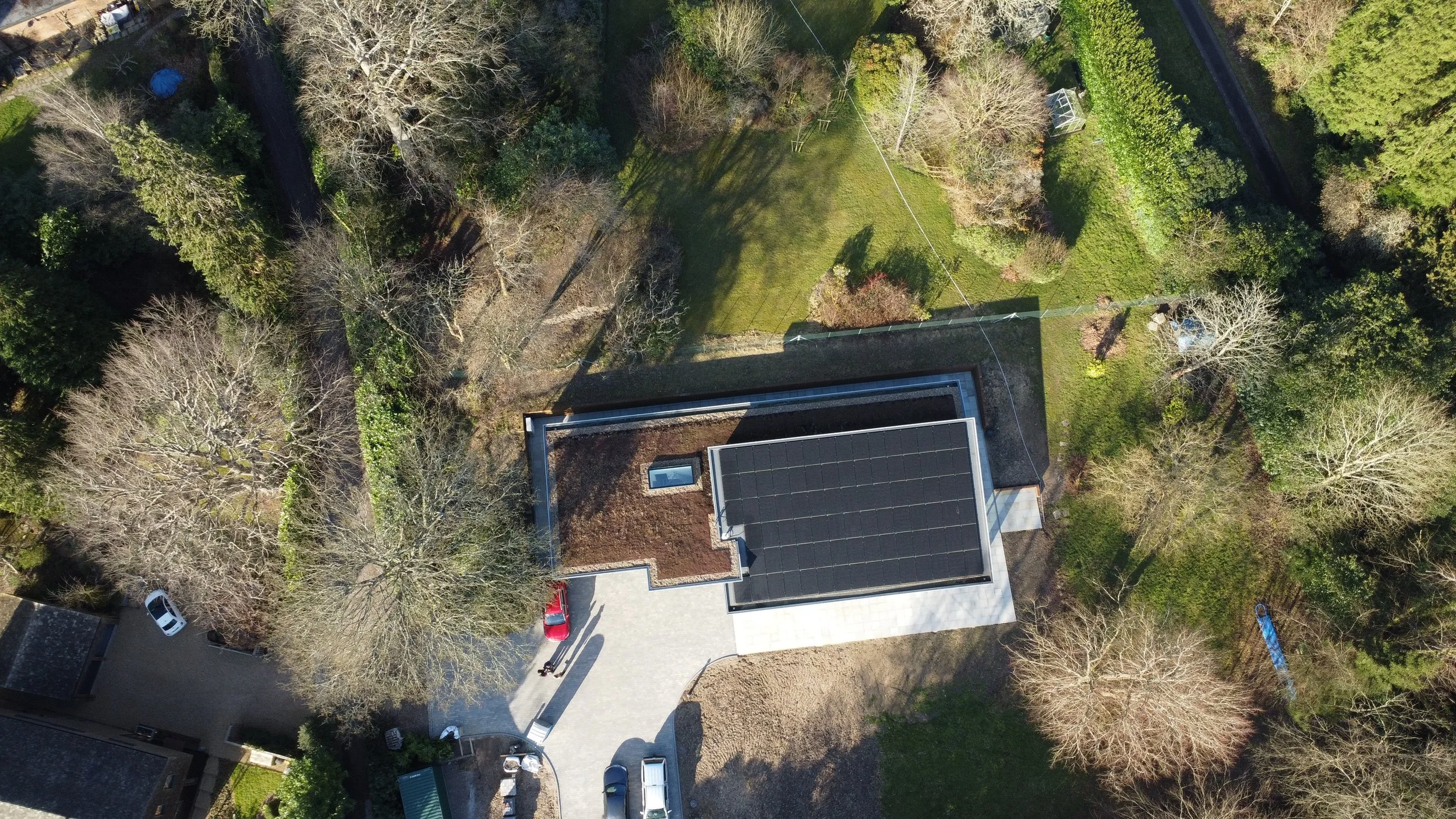 Aerial view of a house with a dark roof covered in solar panels, surrounded by trees and a driveway with cars.