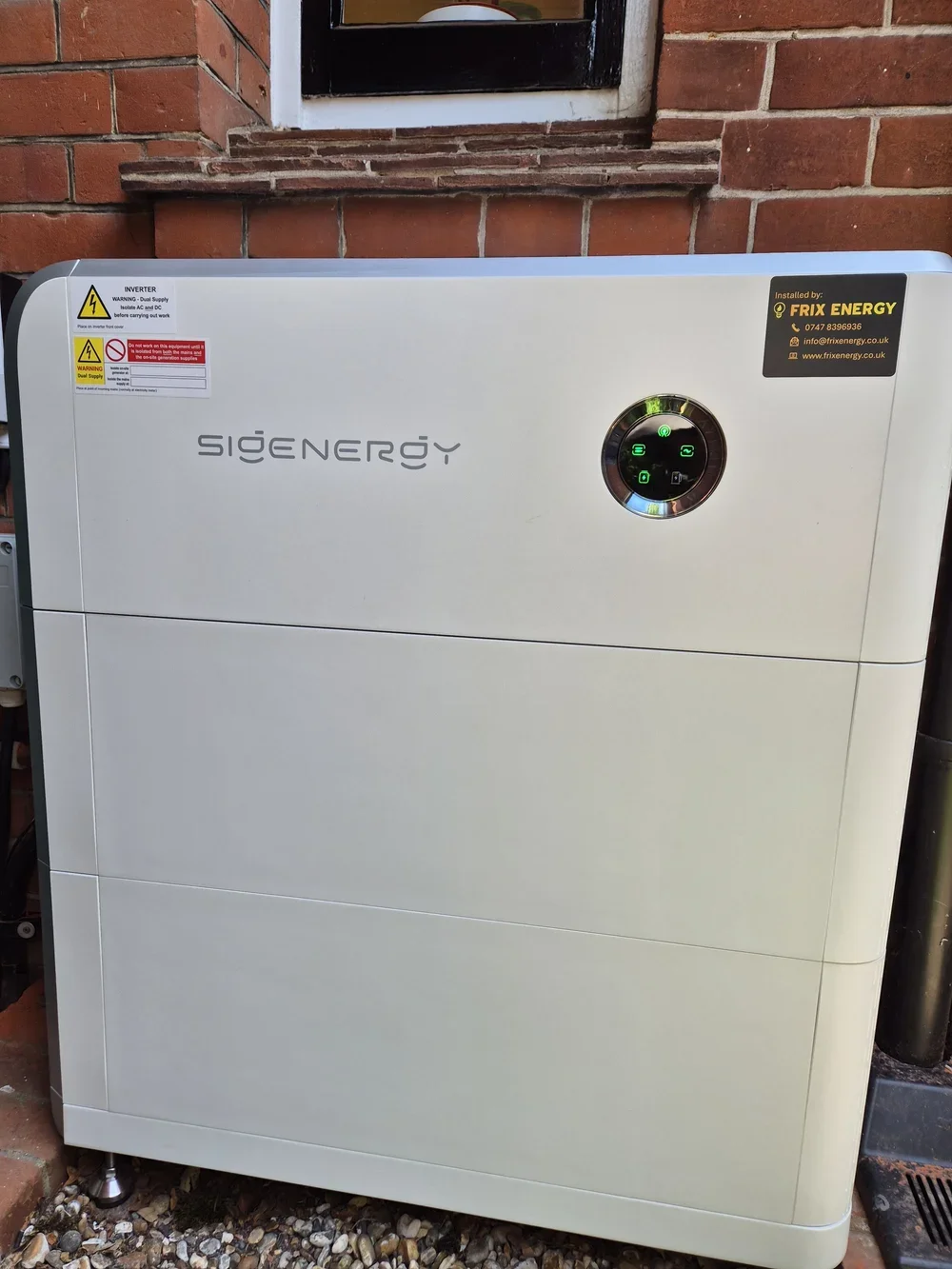 Outdoor white electrical unit with a circular digital display and stickers, attached to a brick wall.