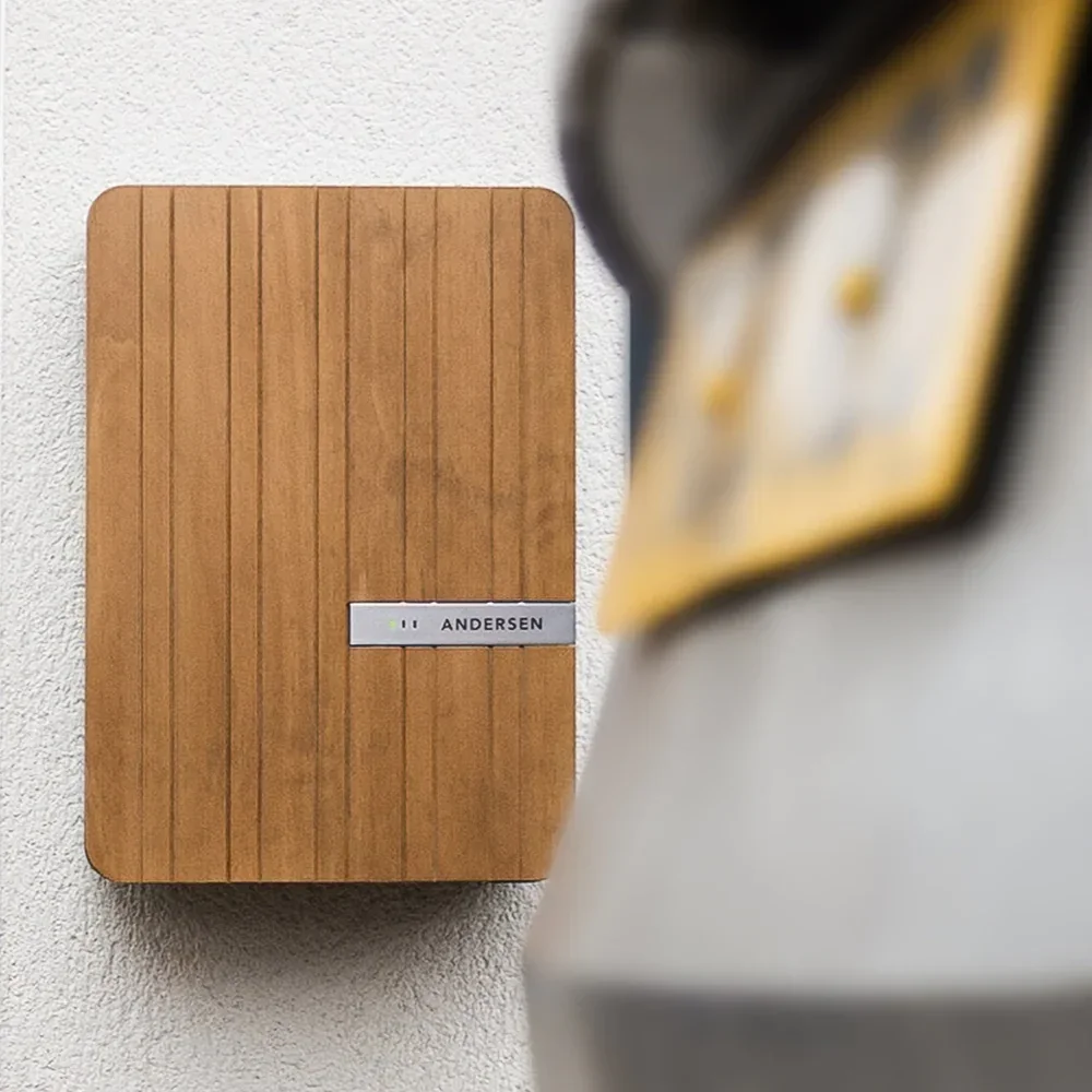 A wooden mailbox with a silver nameplate that reads 'ANDERSEN' is mounted on a white textured wall. Part of a blurred yellow and black object is visible in the foreground.