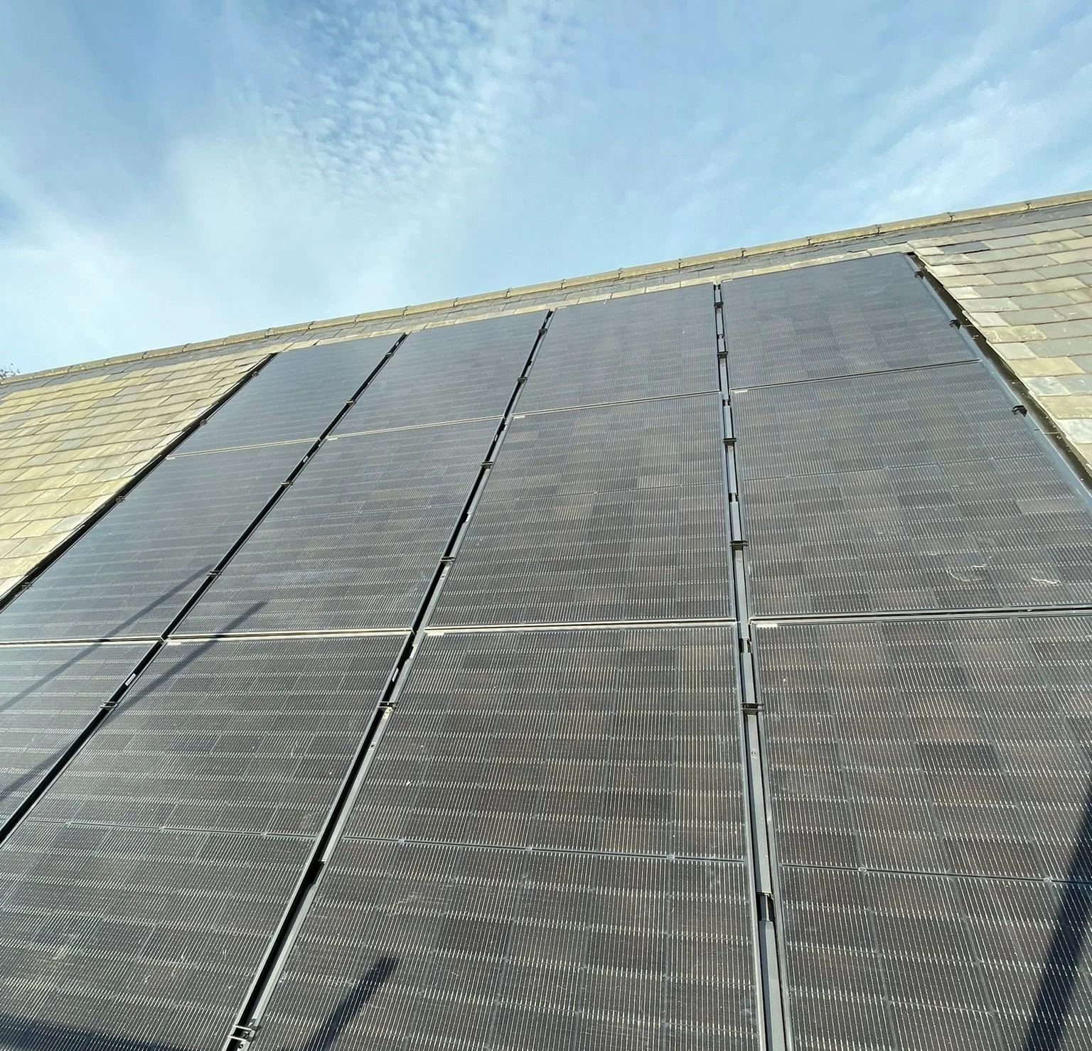 A building with a sloped roof covered with multiple solar panels installed in rows under a blue sky with scattered clouds.