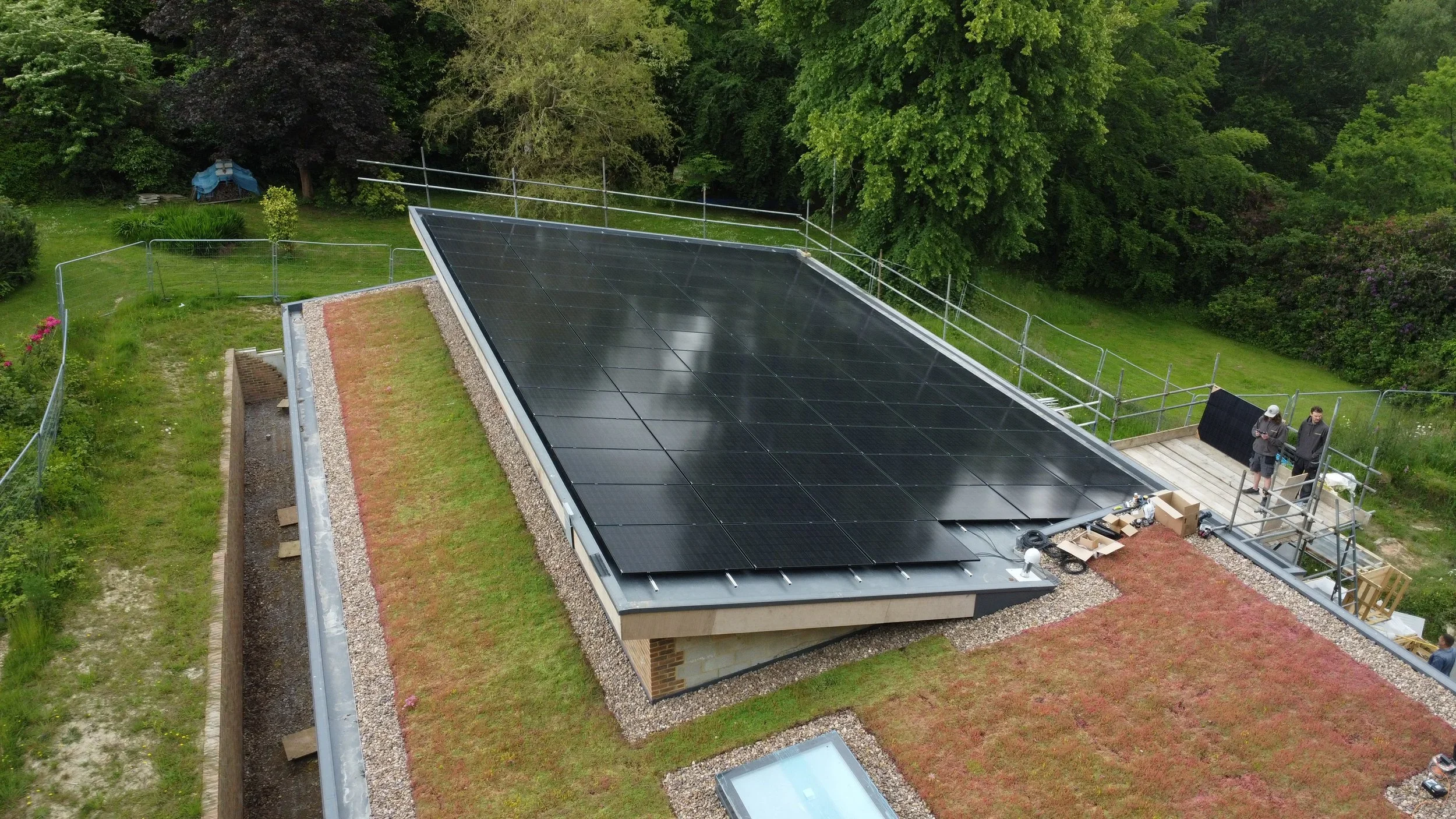 A building with a flat roof covered in solar panels, surrounded by greenery and trees, with scaffolding and two people working on the roof.