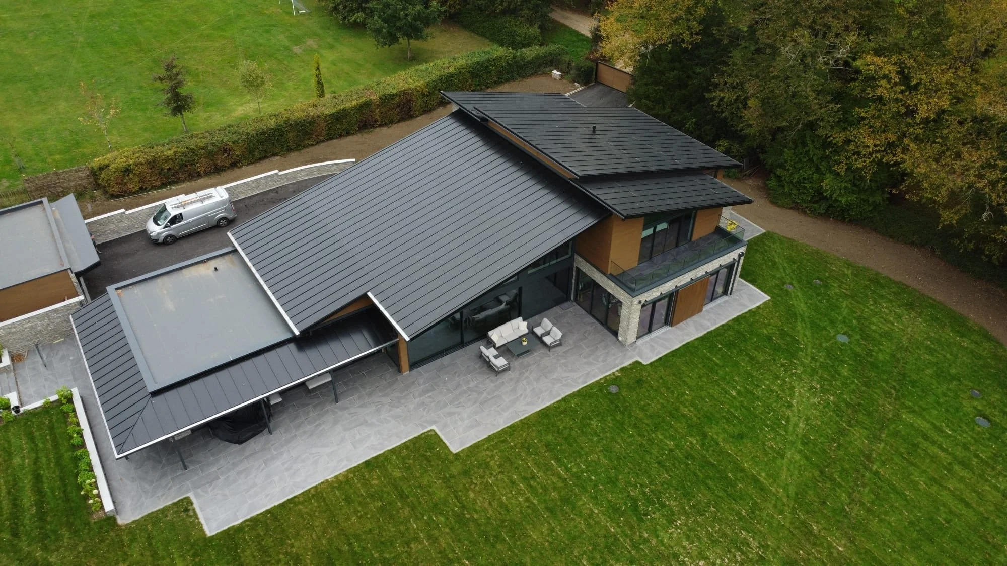 An aerial view of a modern house with a metal roof, outdoor patio furniture, green lawn, trees, and a parked silver van.