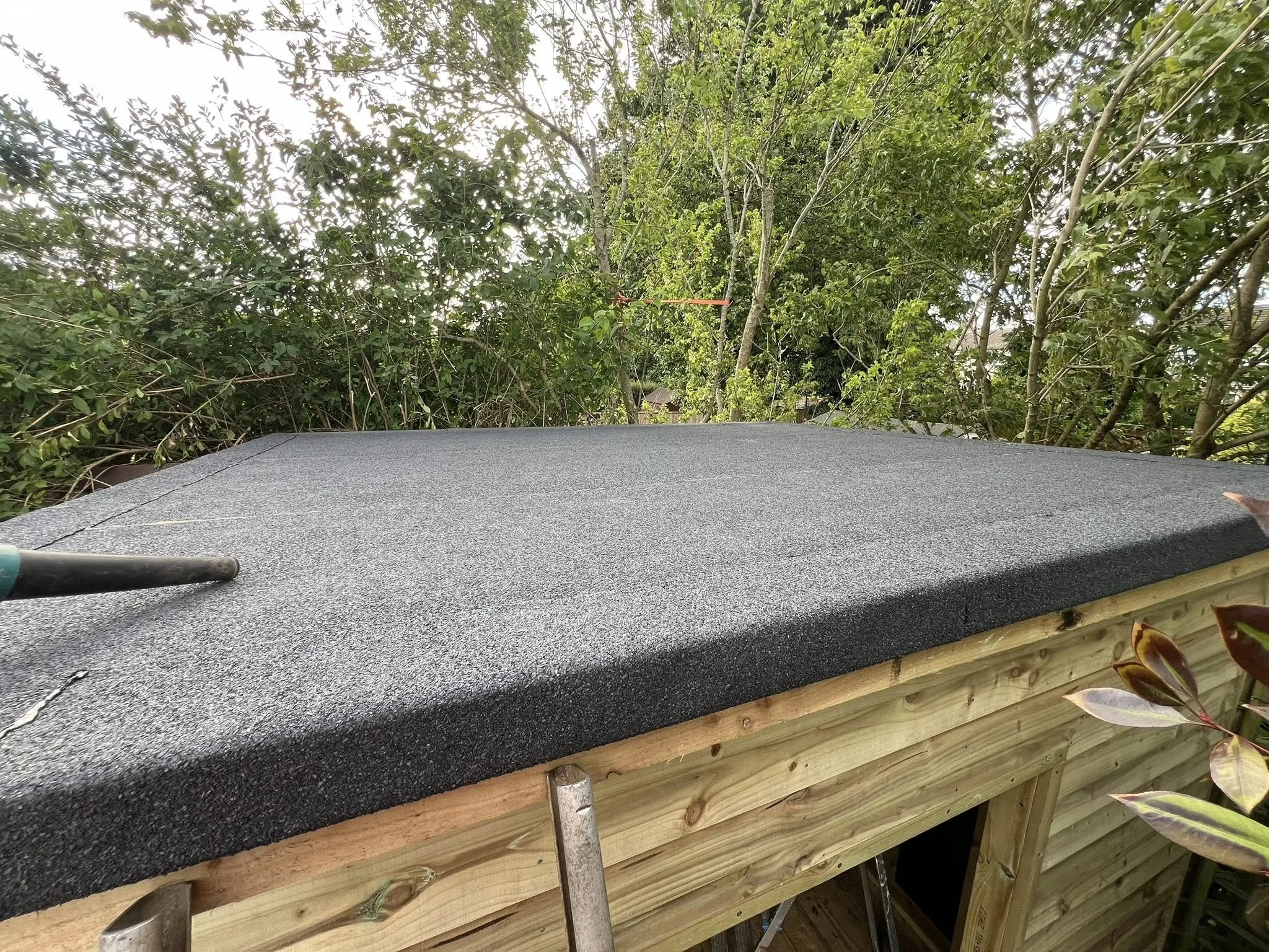 Roof material being installed on a small wooden structure surrounded by trees and greenery, with a screwdriver in the foreground.