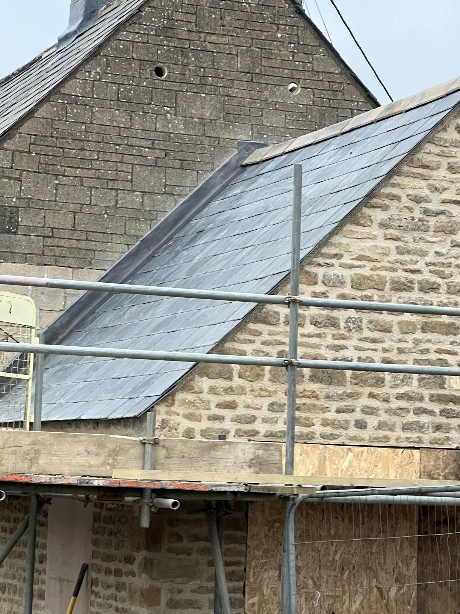 Construction scaffolding and safety railing in front of a stone building with a steep roof covered in slate tiles.