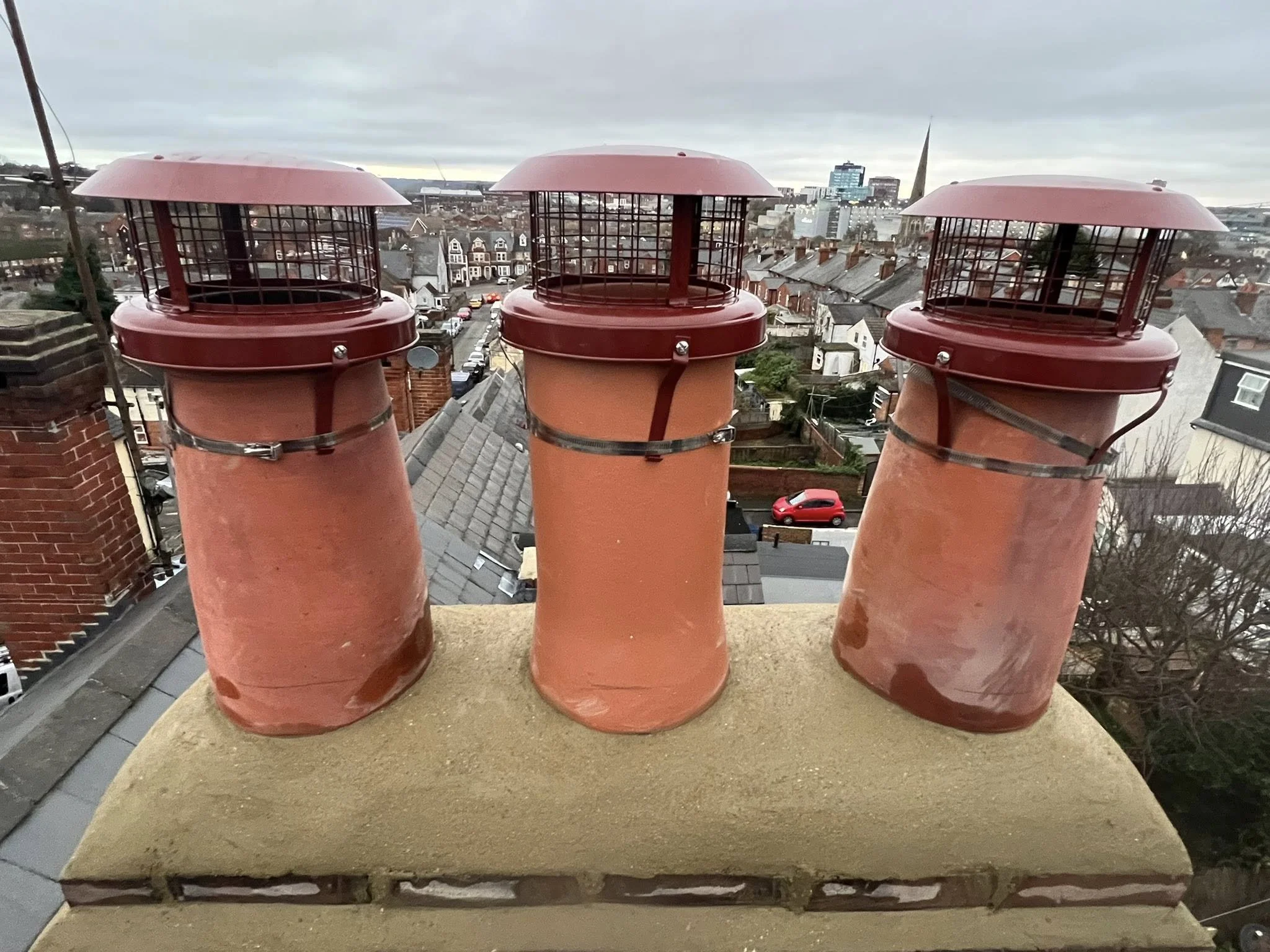 Three red chimney caps with metal wire guards on a rooftop overlooking a cityscape with houses, streets, and taller buildings in the background, under cloudy skies.