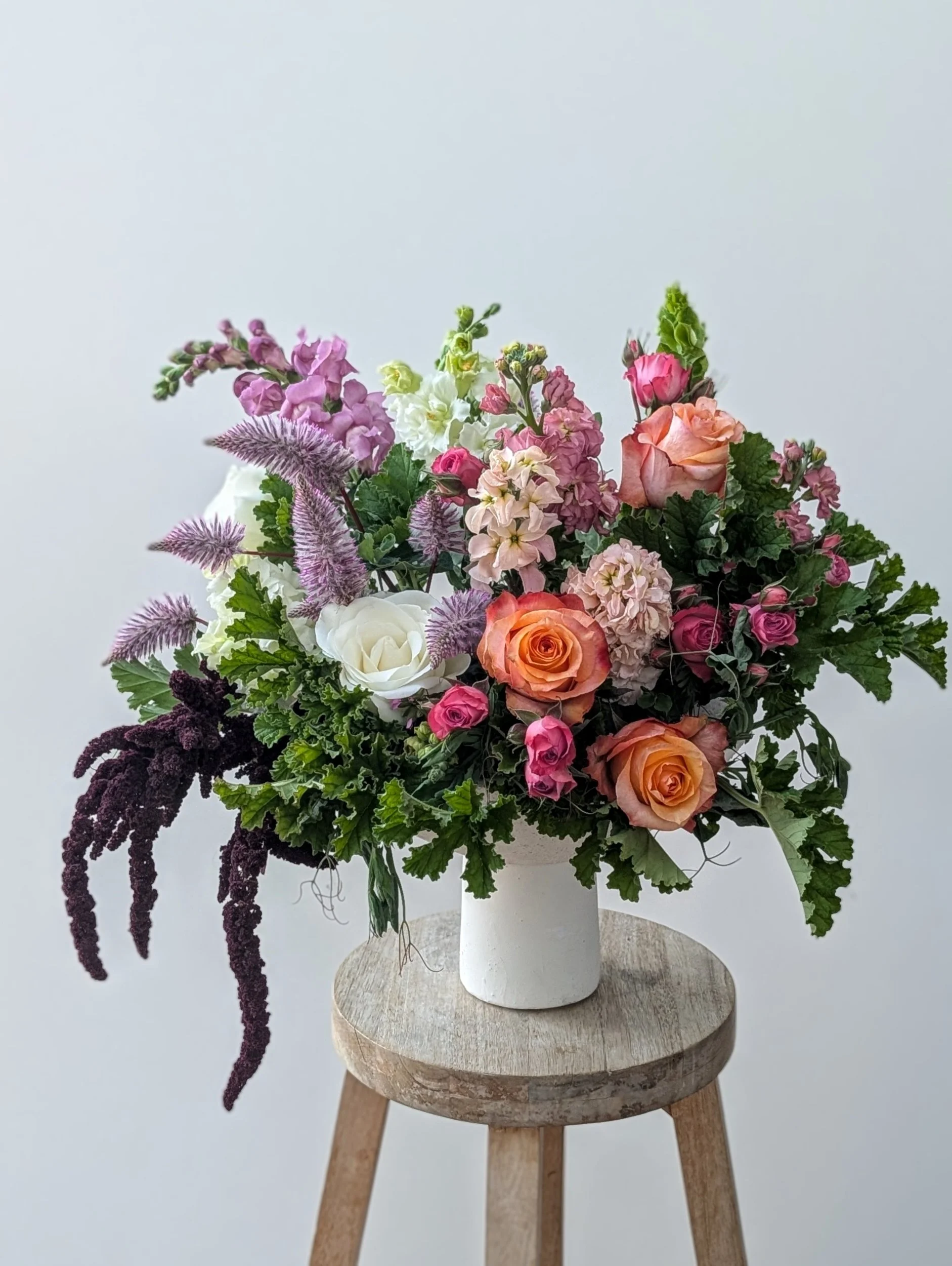 A colorful flower arrangement in a white vase on a wooden stool, featuring roses, hydrangeas, and other mixed flowers.