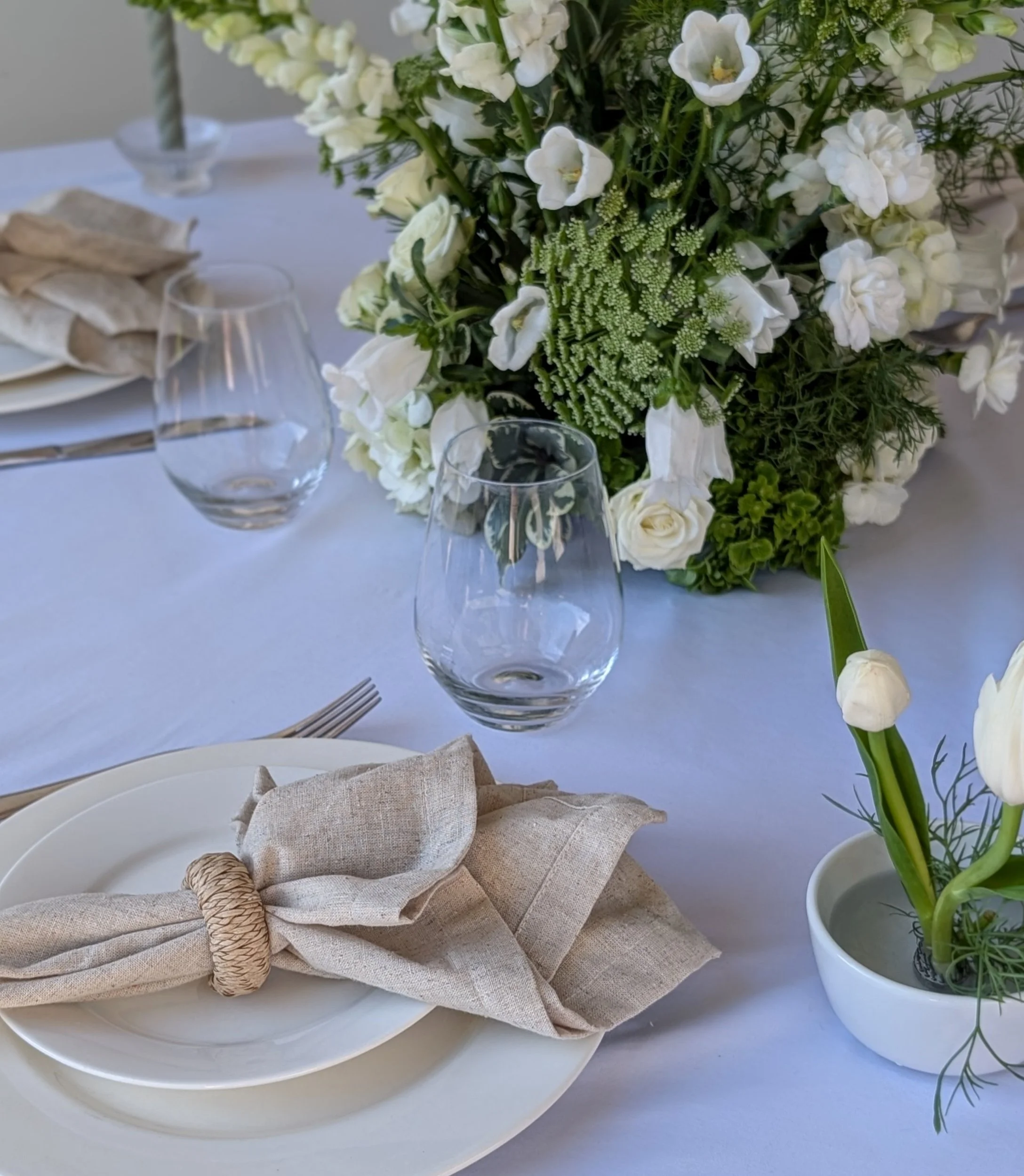 Elegant dining table with a large floral centerpiece of white flowers and greenery, clear wine glasses, beige napkins on white plates, and a small white vase with white tulips.