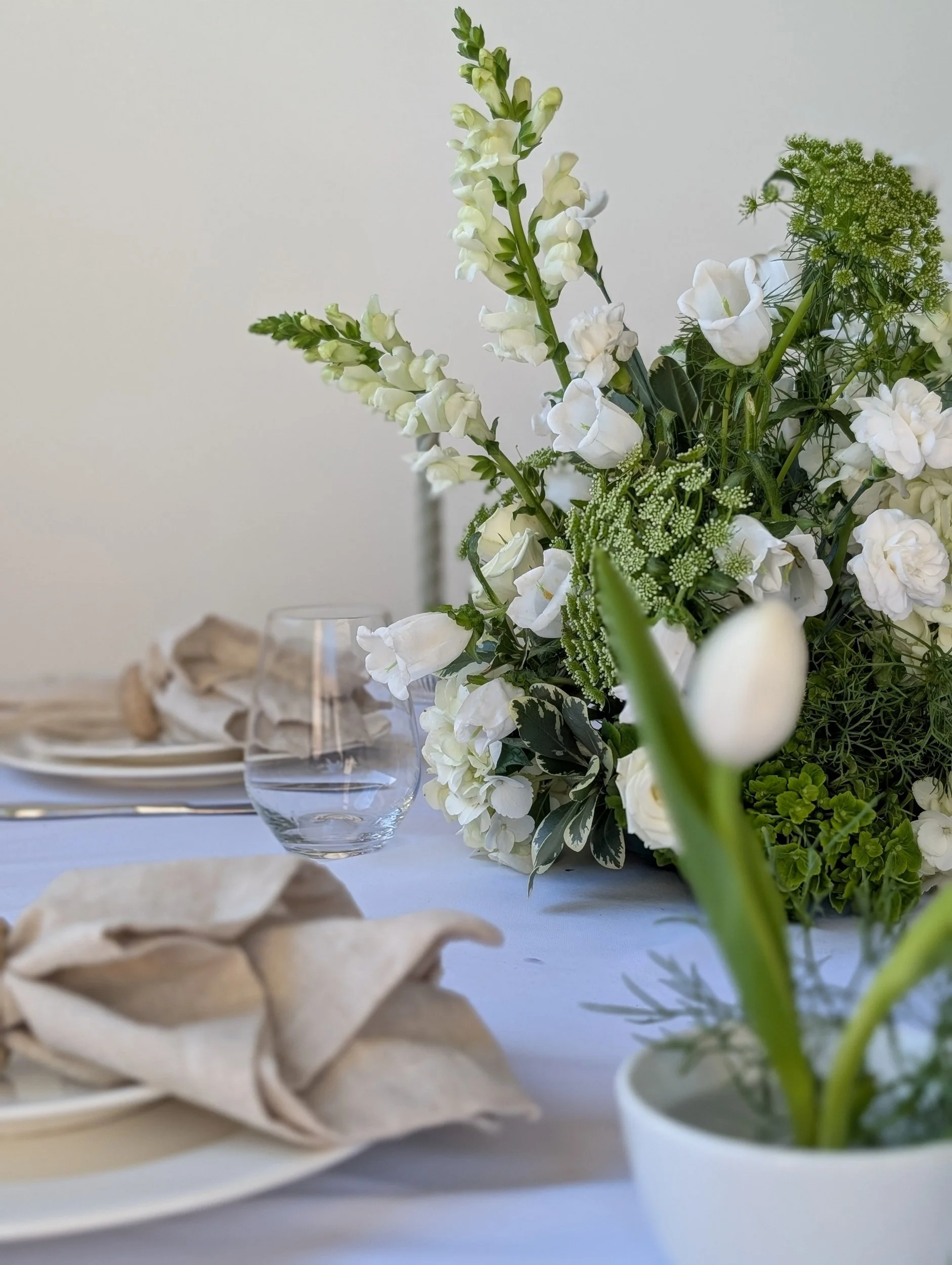 Close-up of a floral centerpiece with white flowers on a dining table with place settings and napkins.