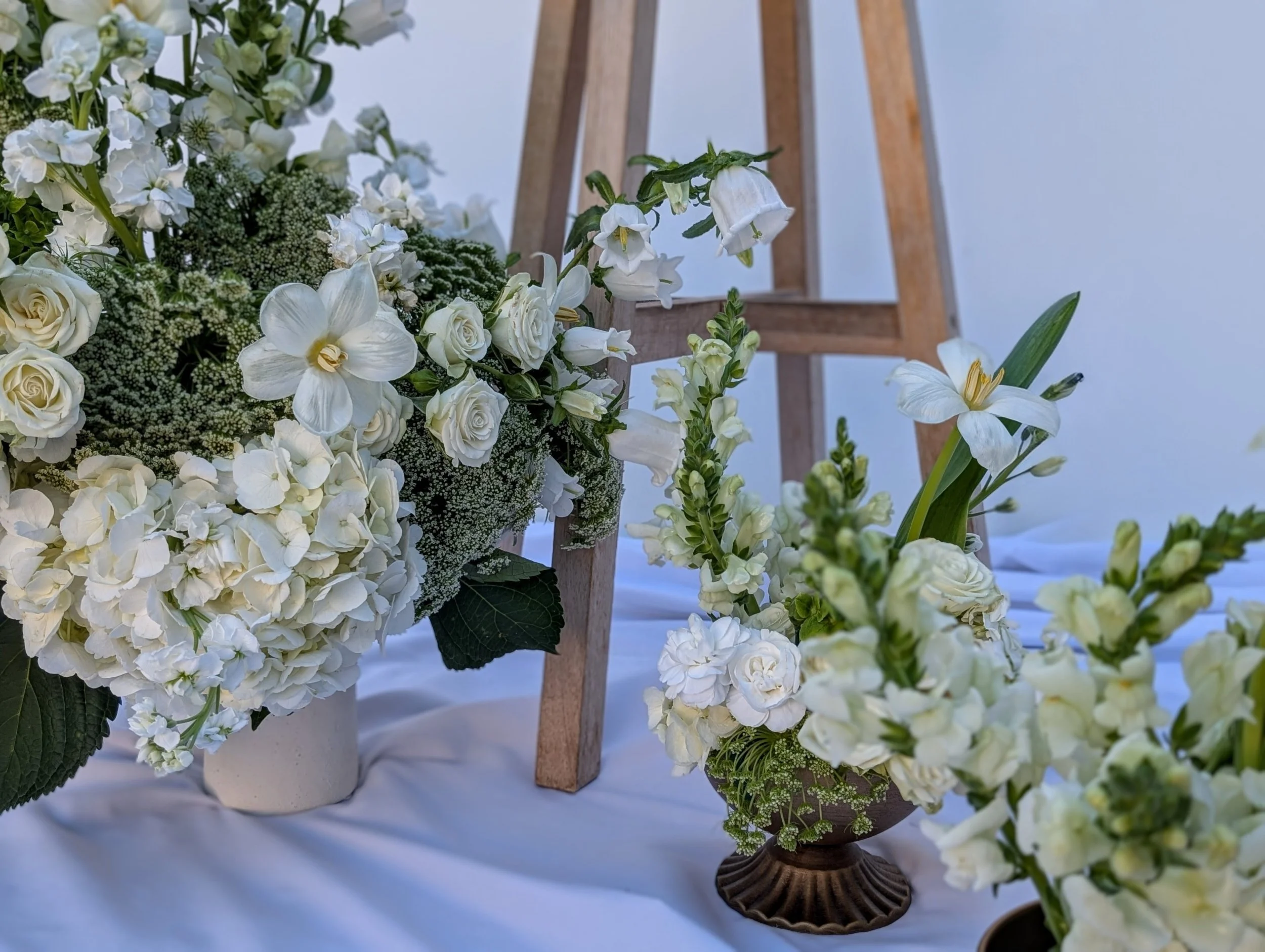 Arranged white flowers including roses, hydrangeas, lilies, and orchids on a white surface against a white and wooden background.