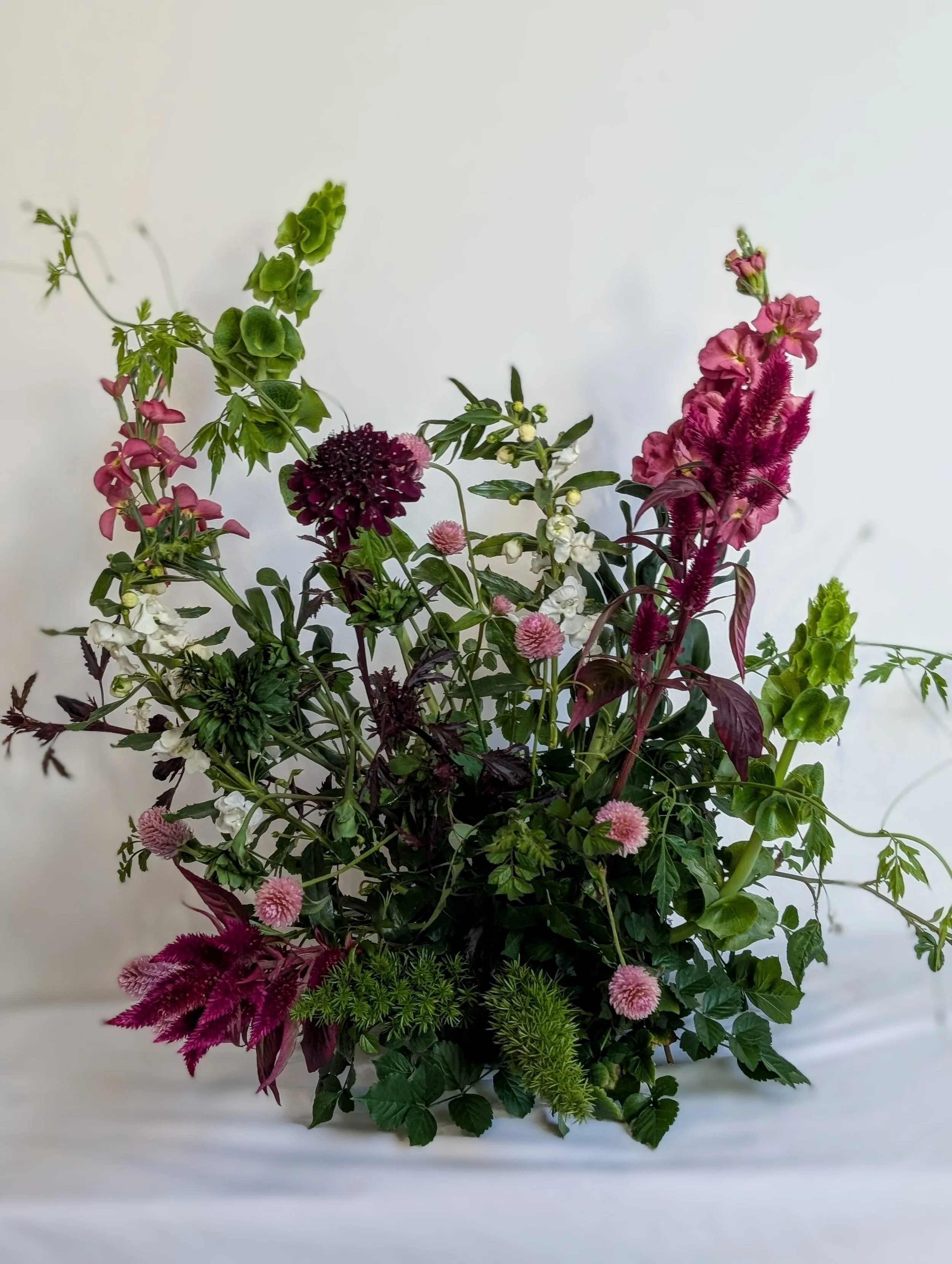A colorful bouquet of mixed flowers and greenery, including pink, purple, and white blossoms against a plain background.