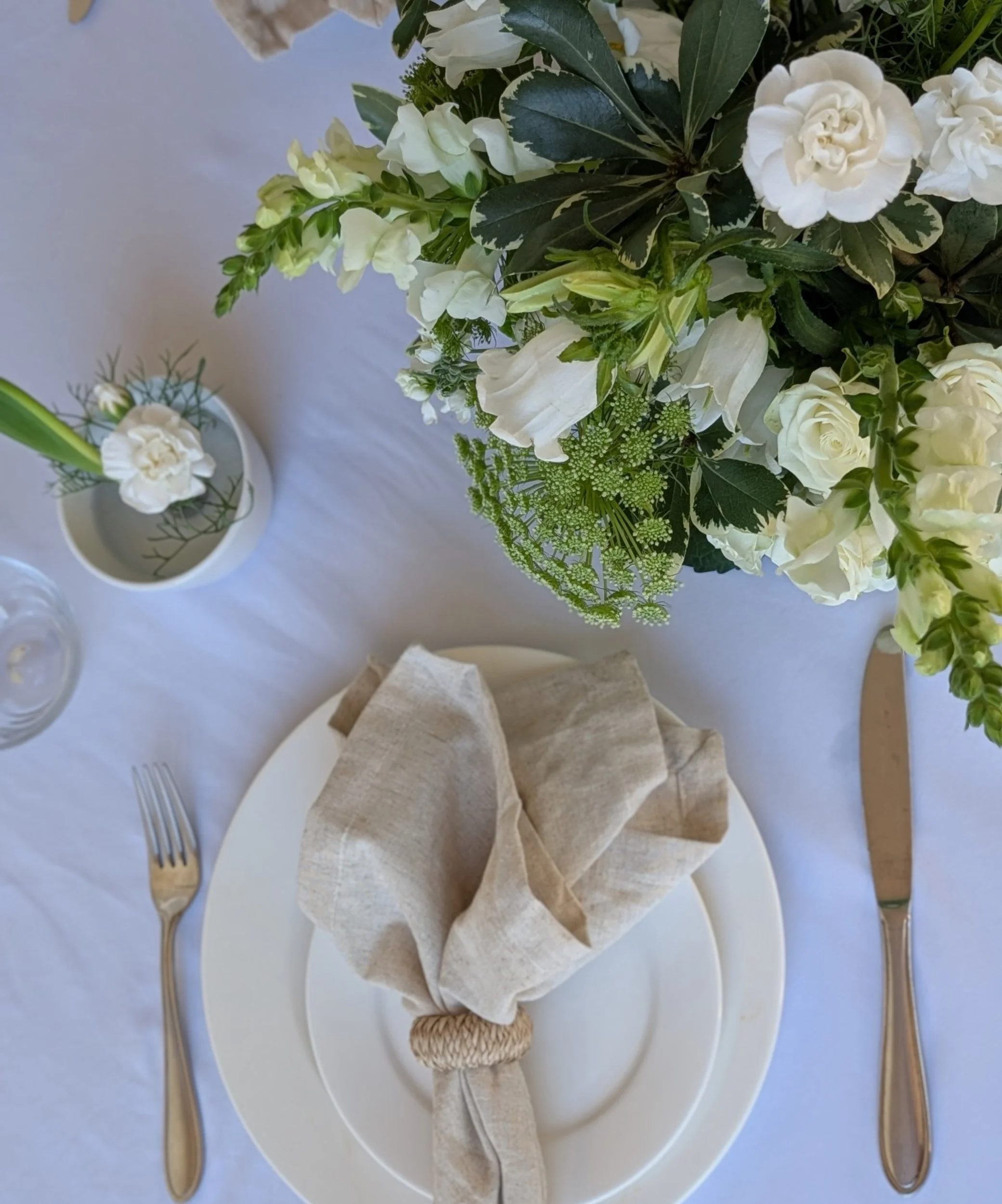 A table setting with a white plate, beige cloth napkin, fork and knife, a small white vase with white flowers, and a large bouquet of white flowers and green foliage.