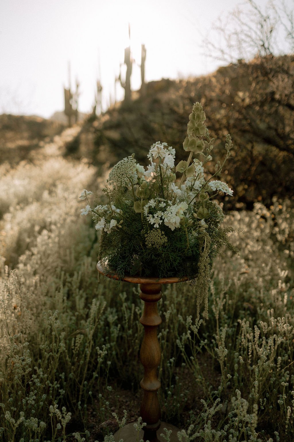 A floral arrangement on a wooden stand in an outdoor desert landscape with cacti and dry bushes, backlit by the sun.