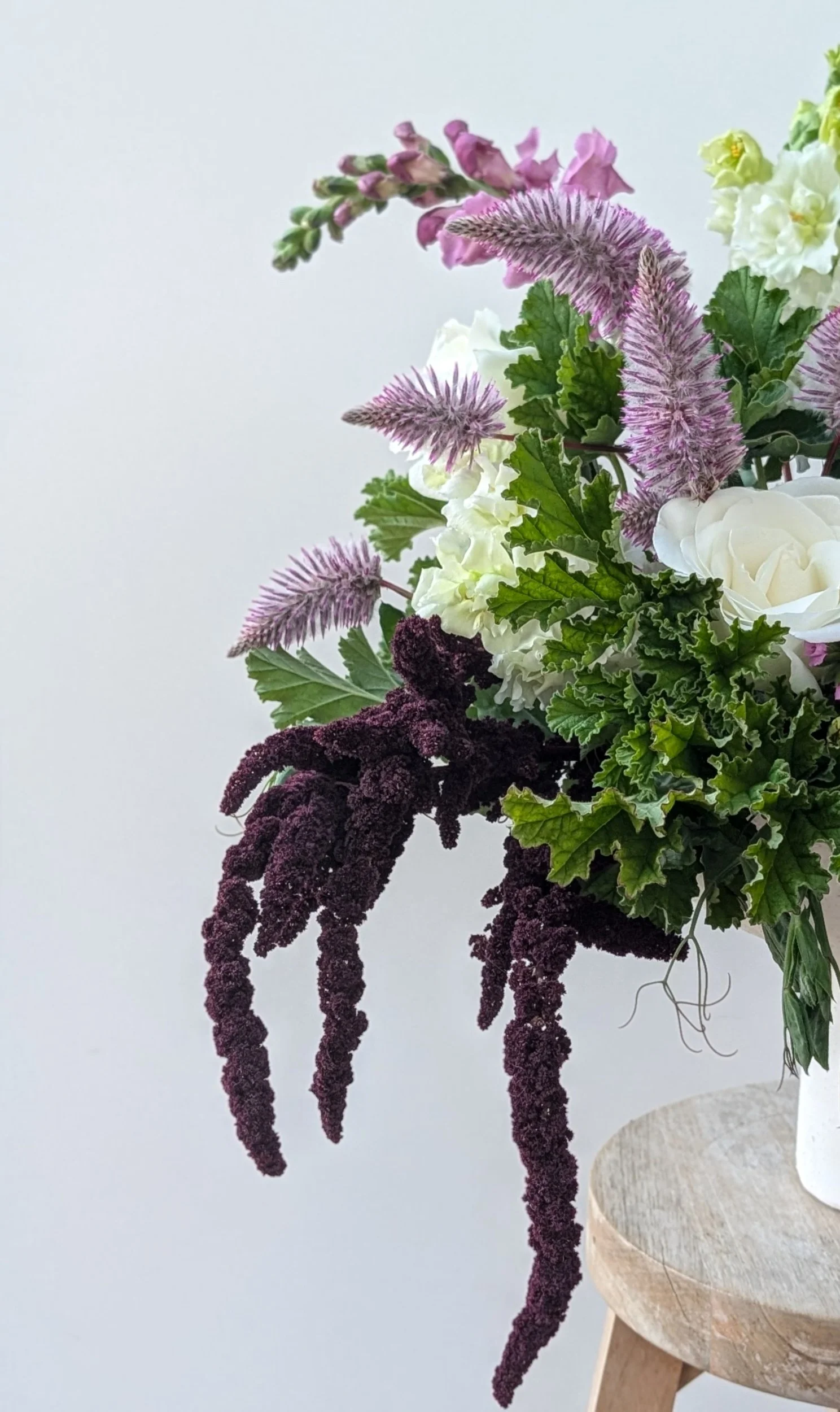 A floral arrangement with white roses, pink and purple flowers, and dark purple cascading amaranthus on a wooden stand against a plain light background.