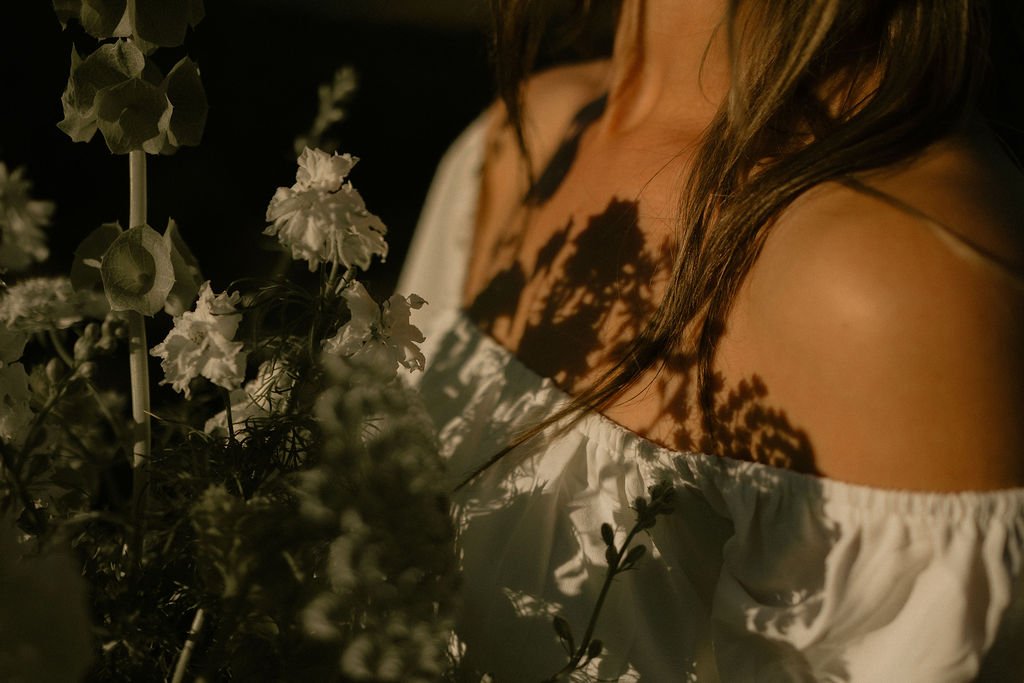 A woman with long hair wearing an off-shoulder white top, with shadows of flowers cast on her skin.
