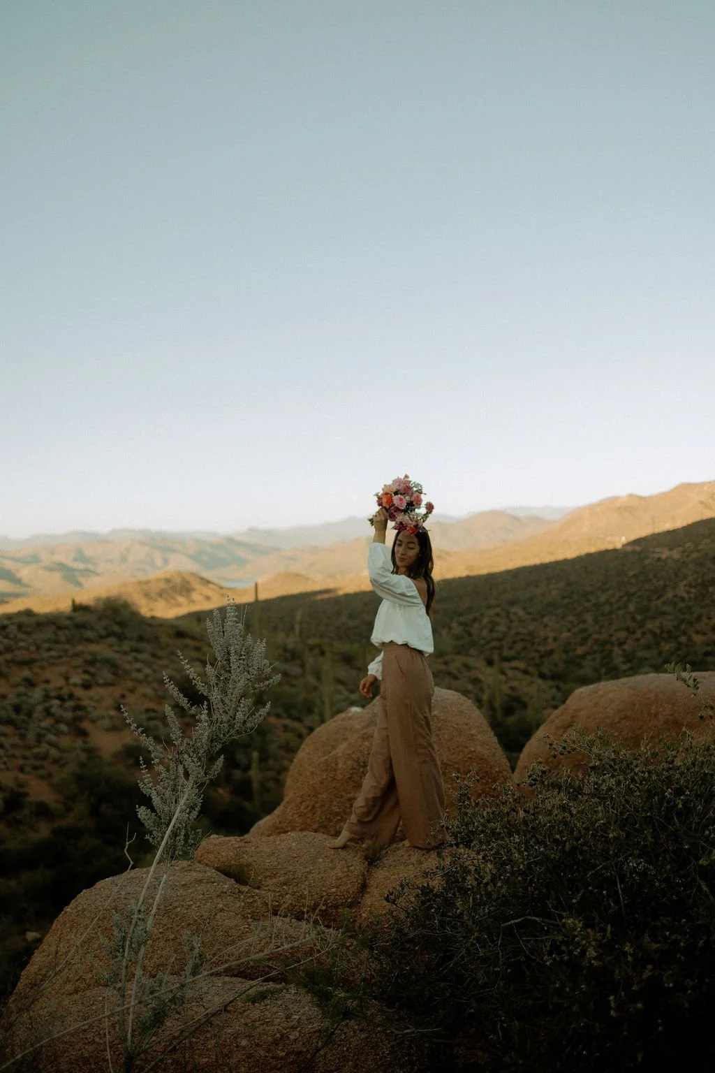 A woman holding a bouquet of flowers above her head while standing on a rocky terrain with desert mountains in the background during sunset.