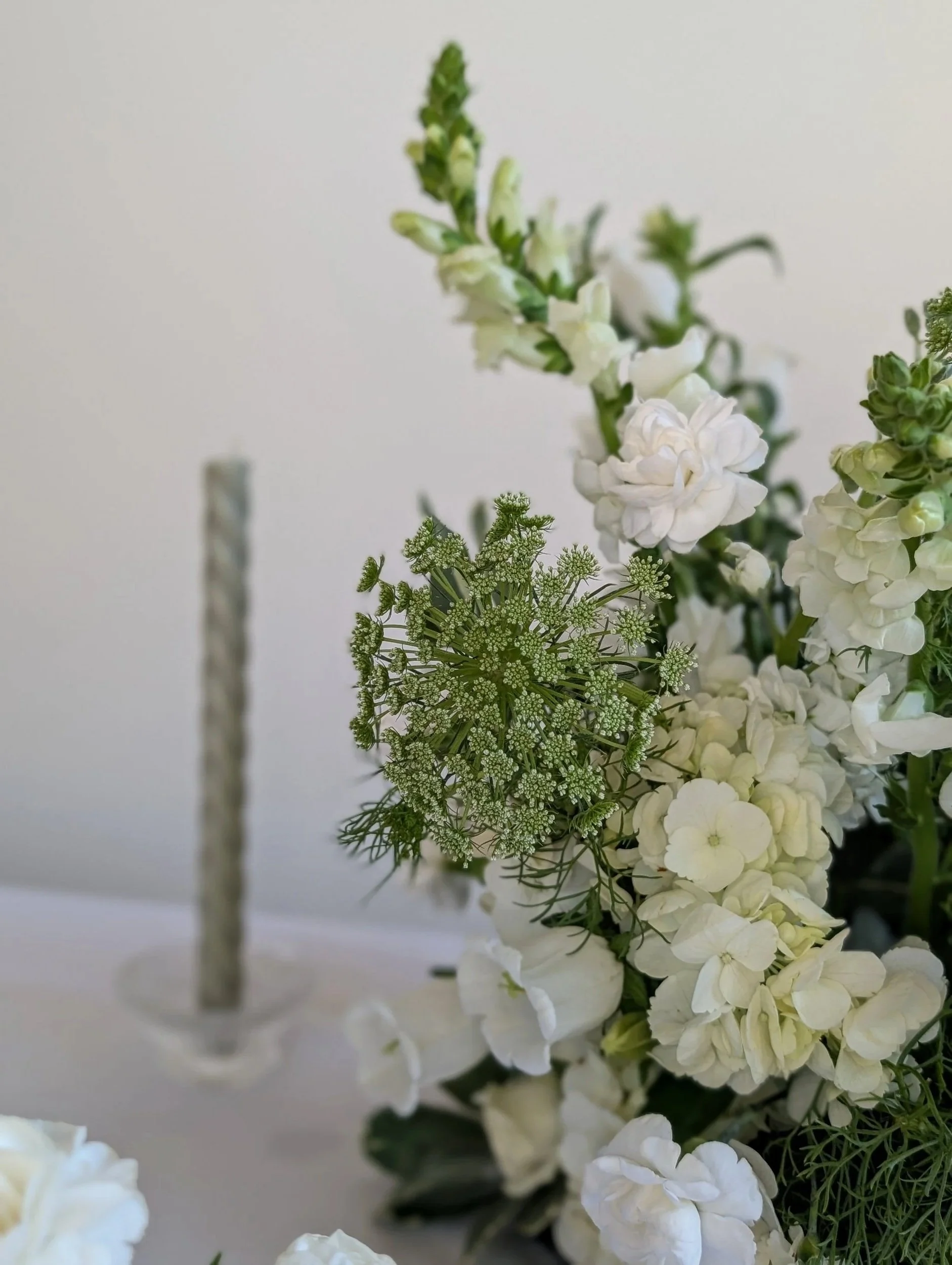 Close-up of a floral arrangement with white flowers, green foliage, and a tall, twisted candle in the background.