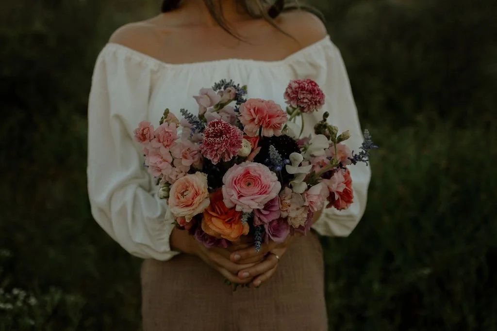 Person wearing a white off-shoulder blouse holding a colorful bouquet of flowers, outdoors.