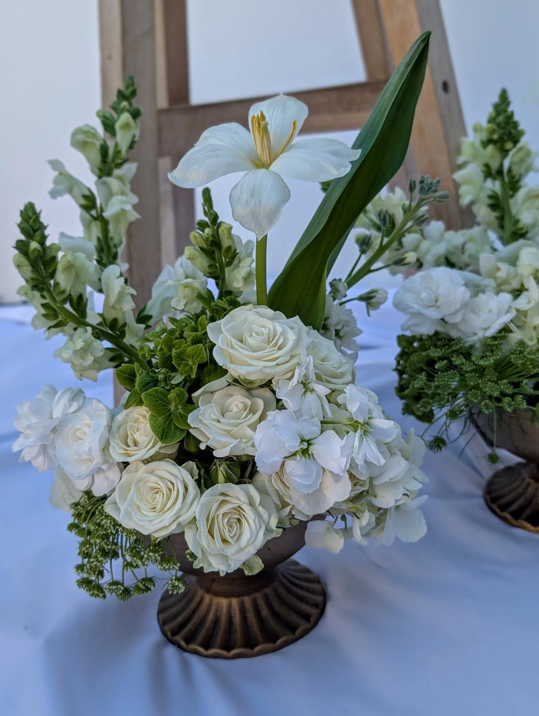 White floral arrangement with roses, a lily, and greenery in a decorative vase on a white table.