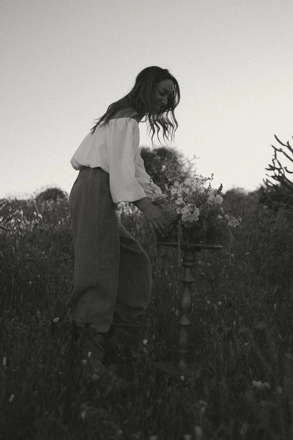 A woman in wide pants and a loose top placing flowers in a small table outdoors at sunset or dusk.