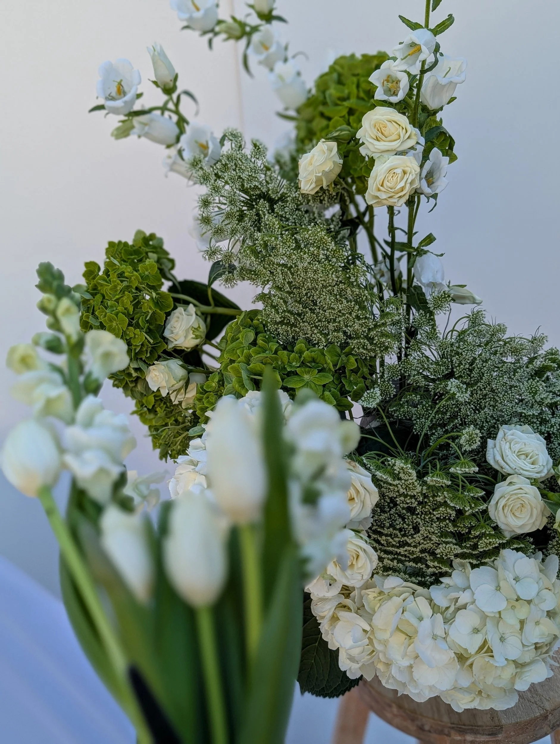 A bouquet of white flowers including roses, tulips, and hydrangeas, along with green foliage and white filler flowers, arranged in a rustic container.
