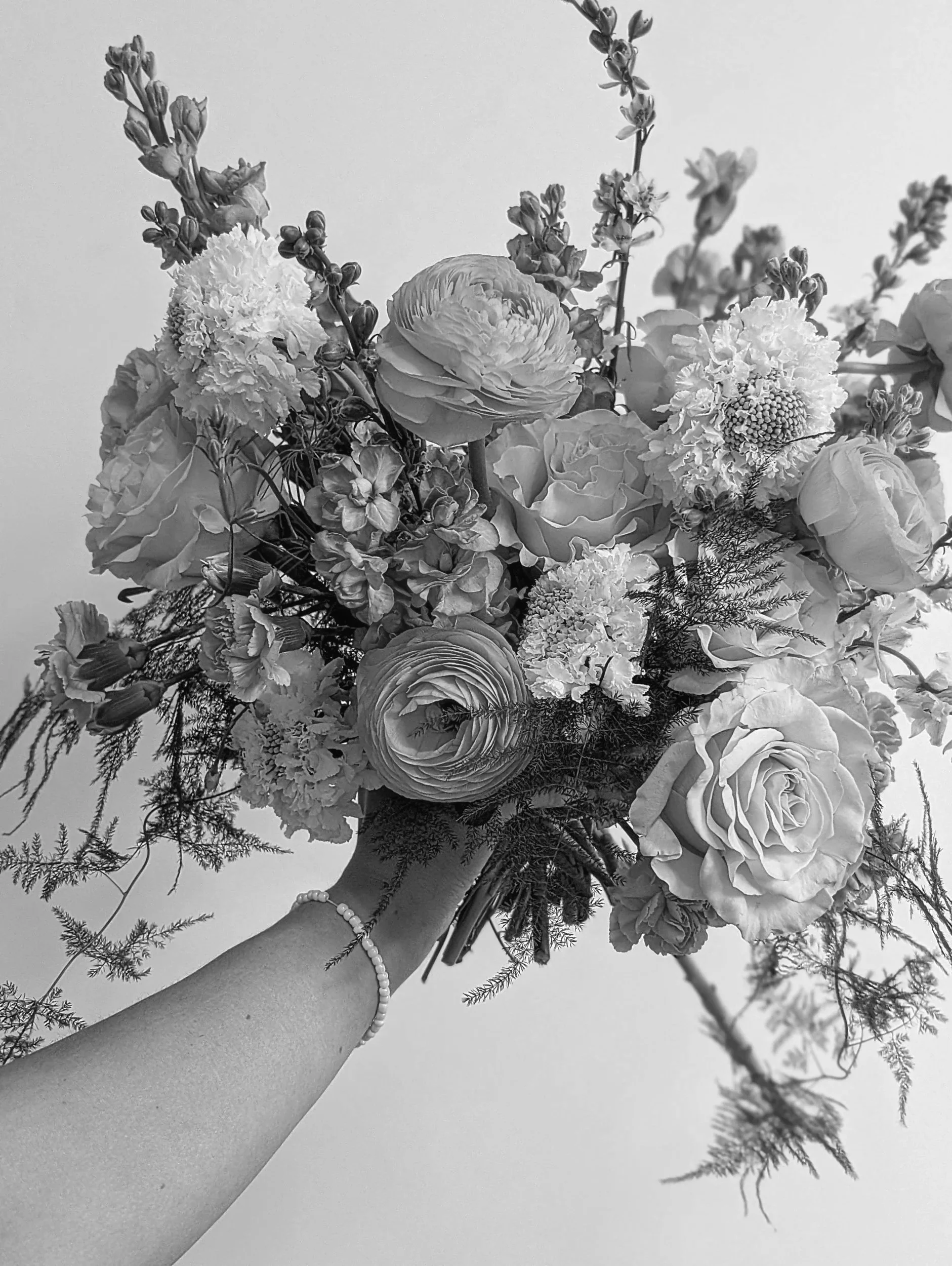 A person holding a bouquet of various flowers including roses, ranunculus, and other blossoms, with some greenery, in black and white.