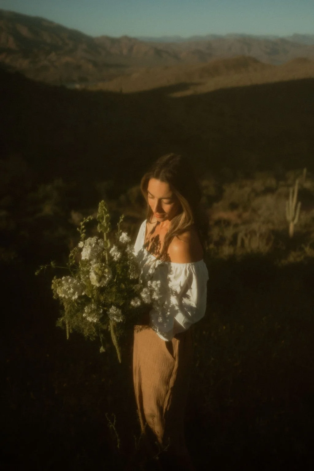 A woman holding a large bouquet of white flowers in a desert landscape during sunset.