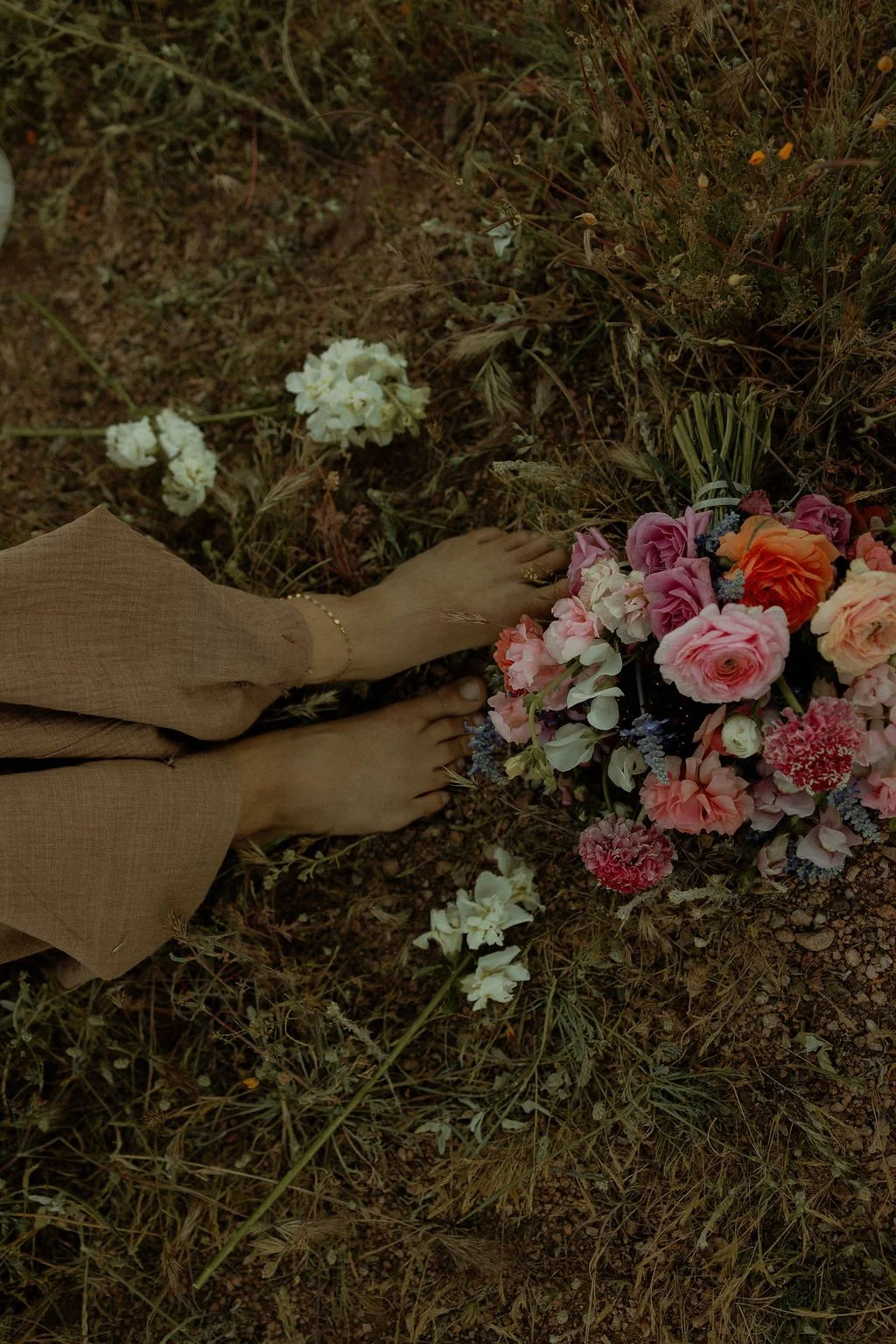 Person with bare feet and beige clothing lying on the ground next to a colorful bouquet of flowers, in a natural outdoor setting with grass and small plants.