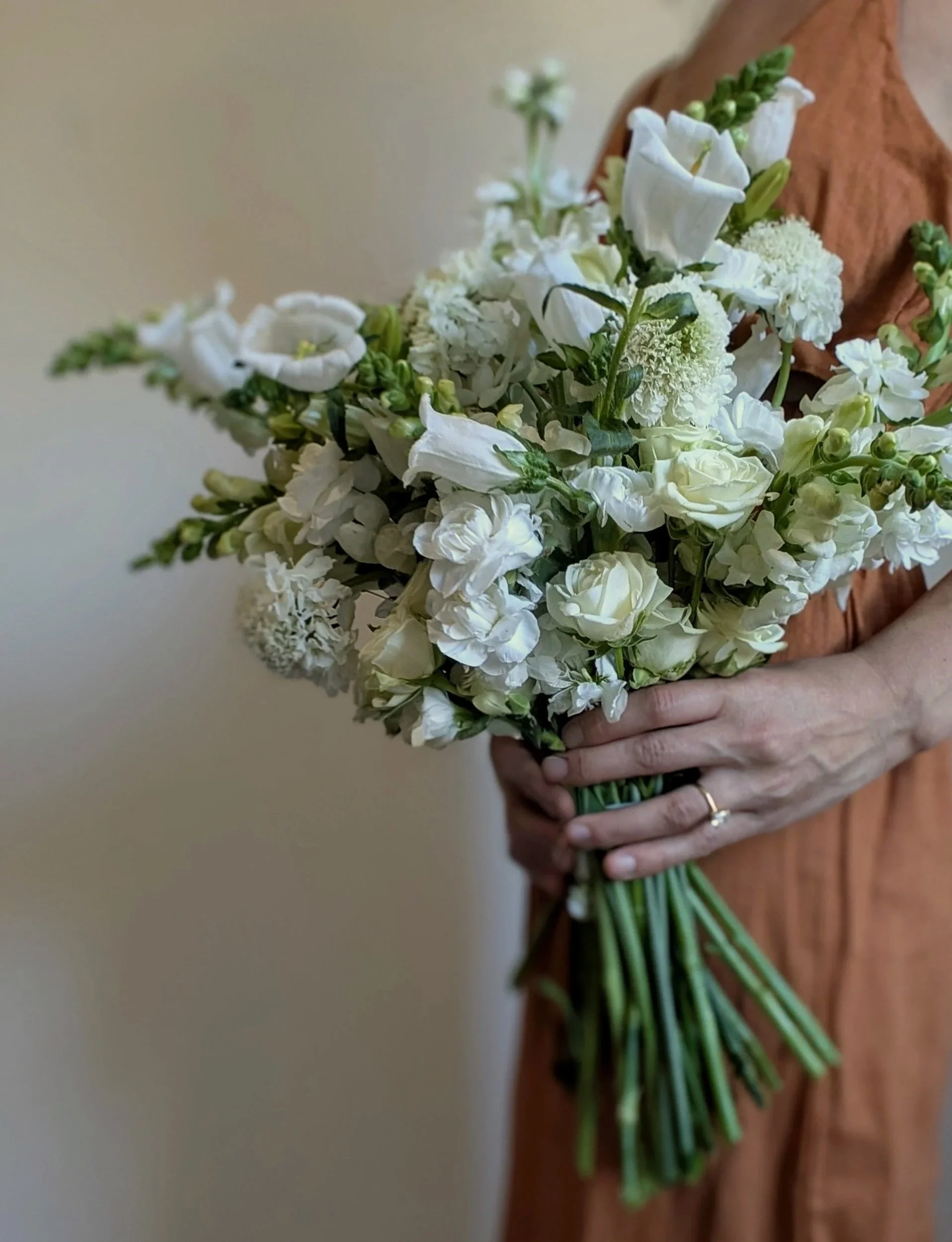 A woman in an orange dress holding a bouquet of white flowers, including roses, lilies, and other blooms, with a ring on her finger.