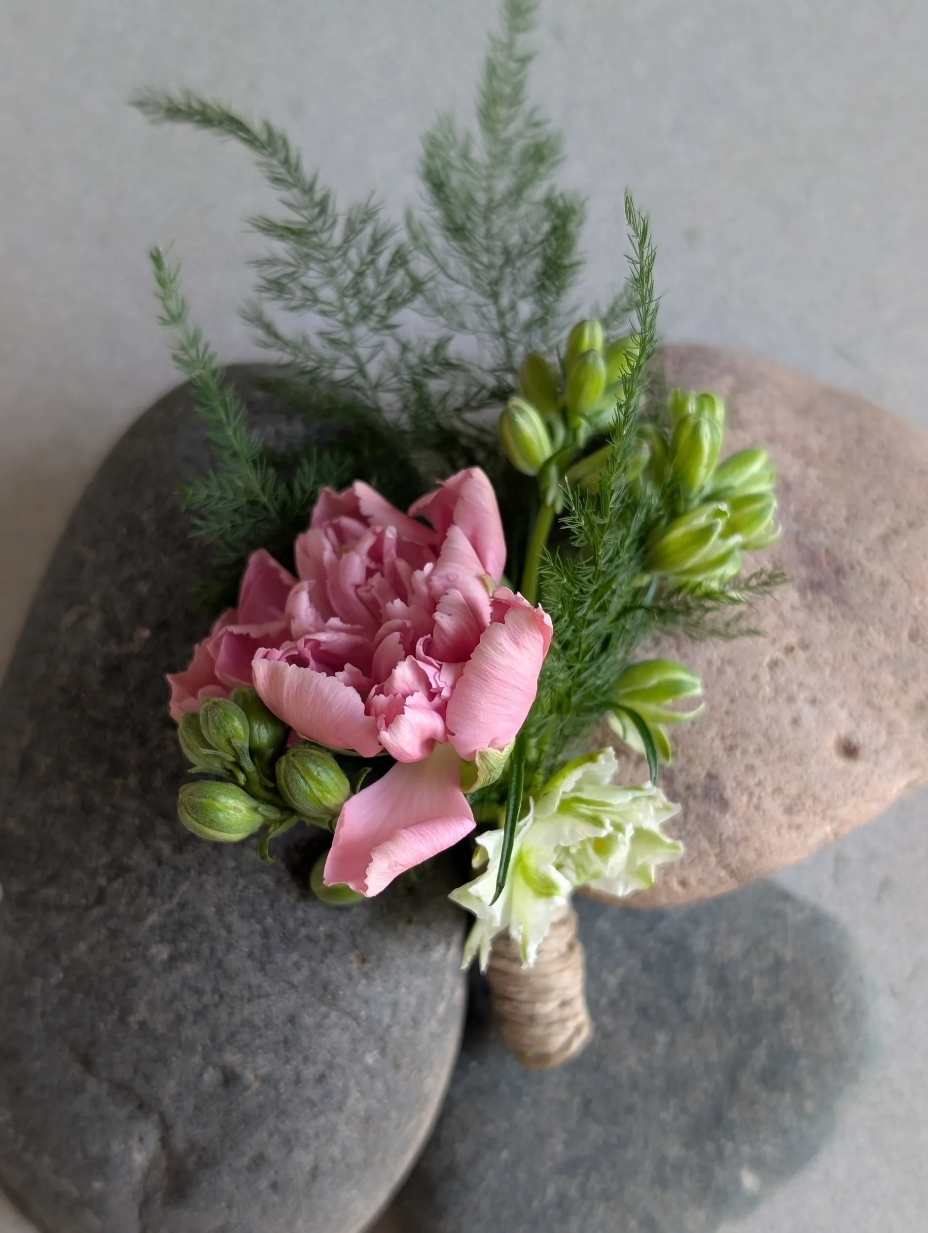 Small flower arrangement with pink and white flowers, green buds, and greenery, wrapped with twine, placed on large gray rocks.