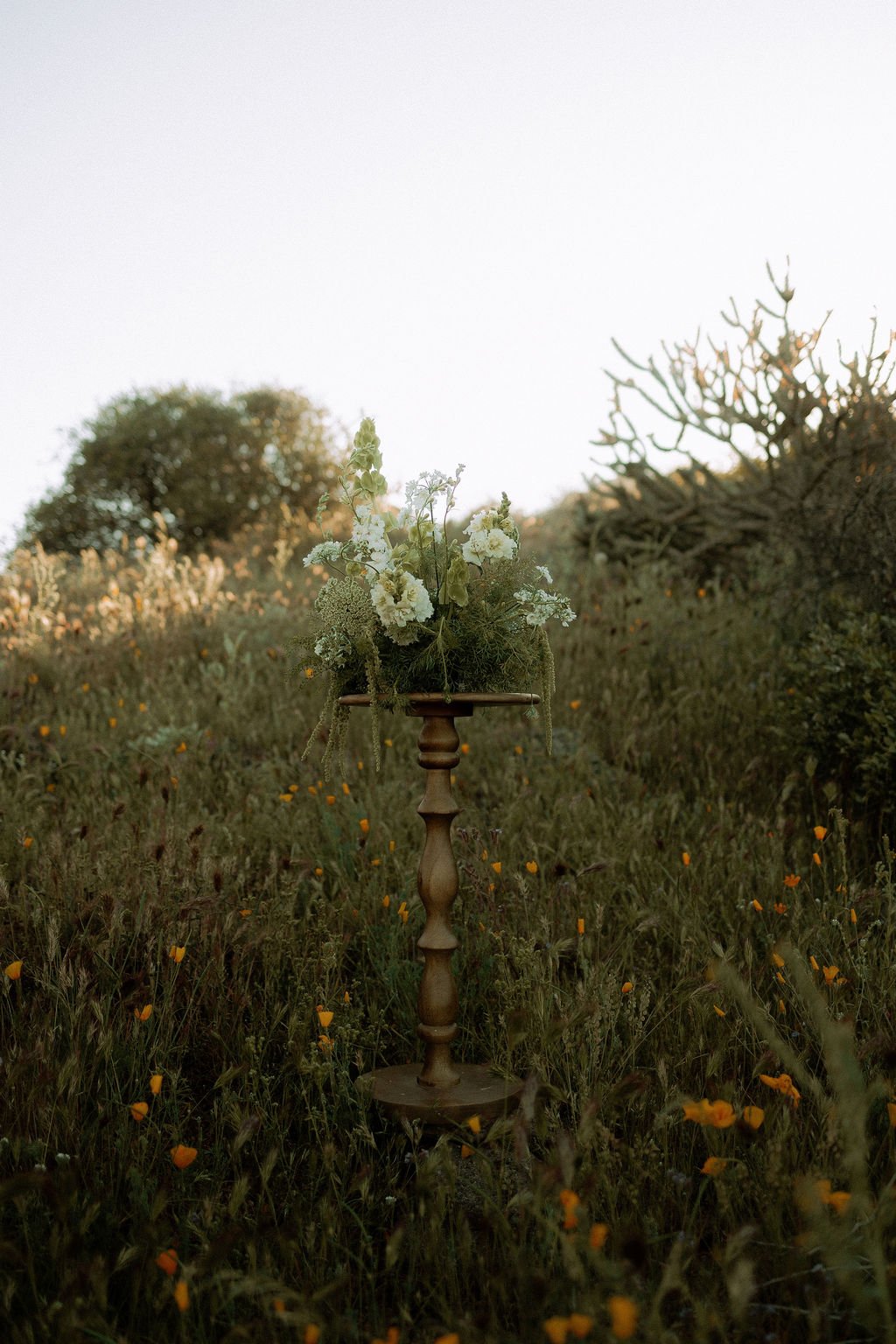 A floral arrangement on a wooden stand in a field with yellow flowers and trees in the background during sunset.