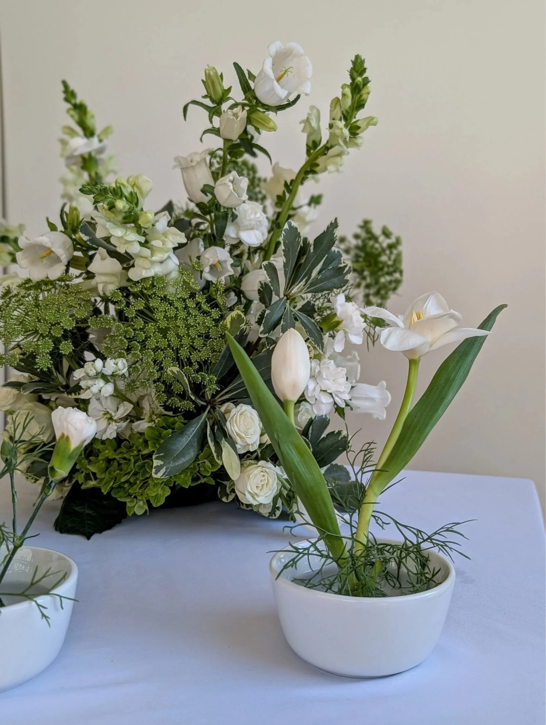 White tulips and other white flowers in a small white pot, with a larger floral arrangement in the background.