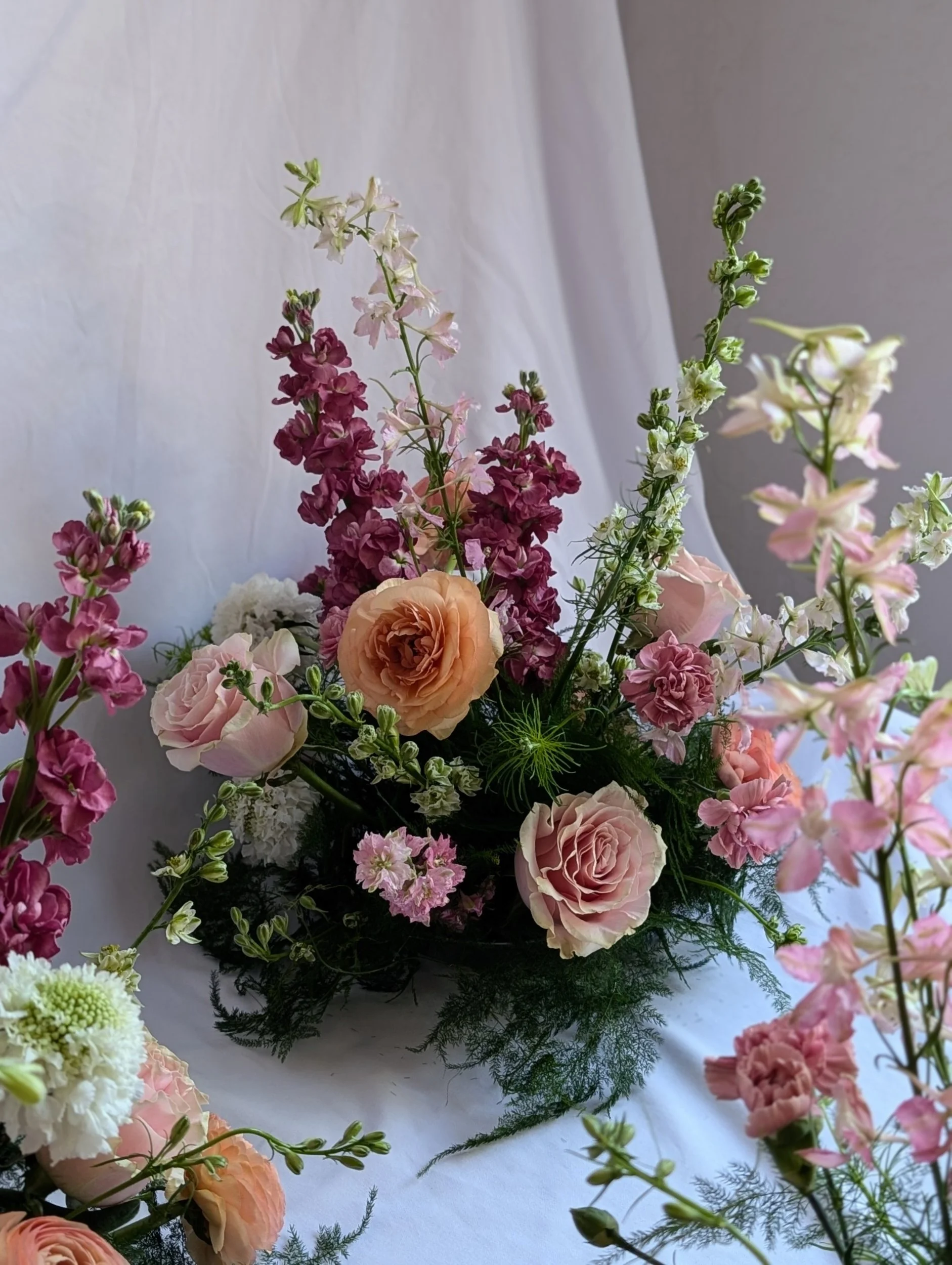 A floral arrangement with pink roses, purple and white stock flowers, and greenery, set on a white surface with a white and gray background.