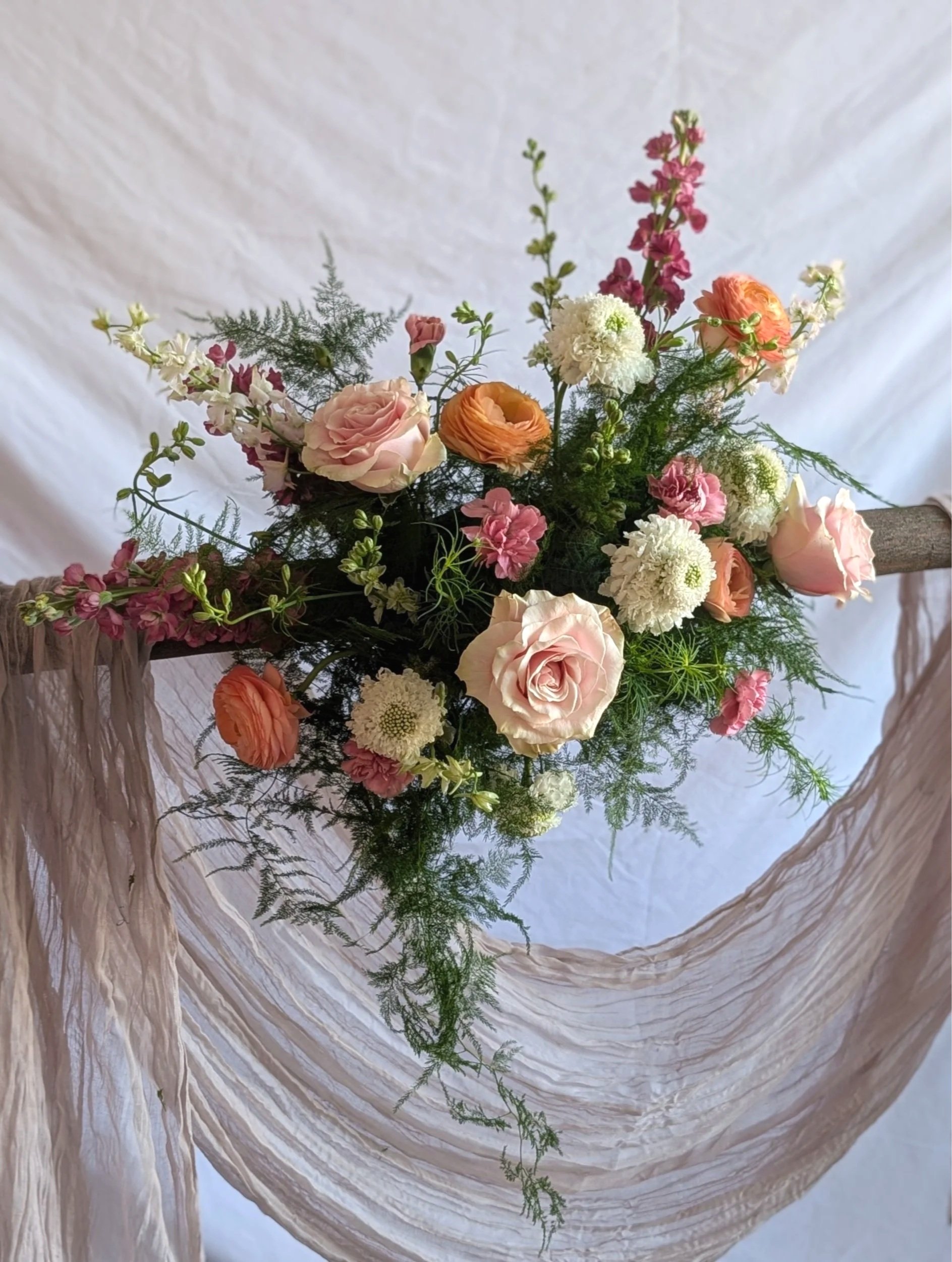 A floral arrangement with pink, white, and orange flowers, greenery, and a draped beige cloth background.