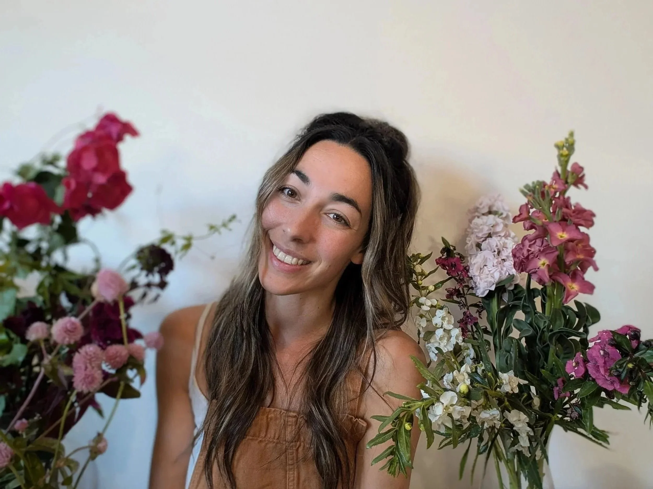 A woman with long brown hair smiling, surrounded by pink, white, and purple flowers.