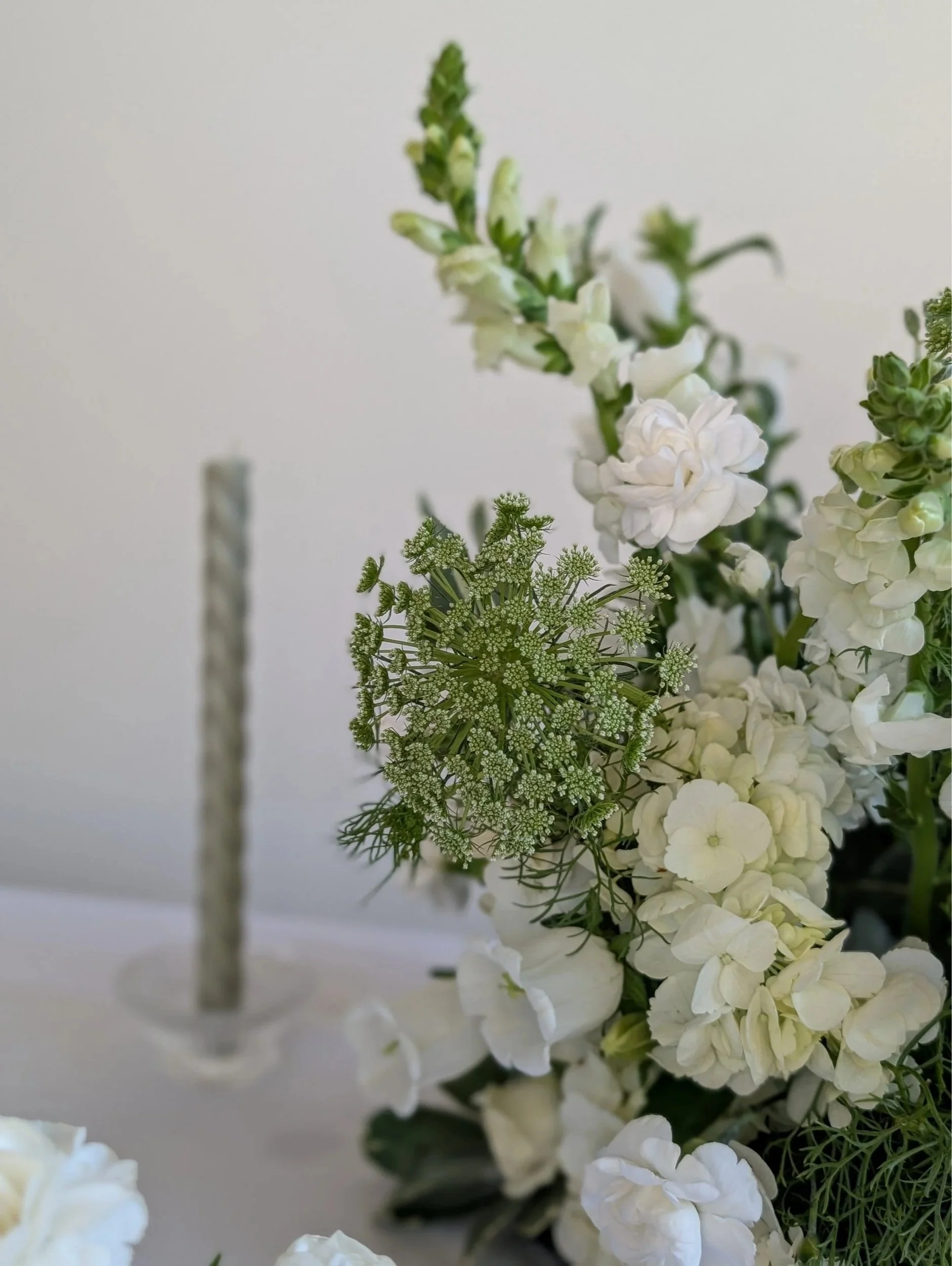 Close-up of white flowers and green foliage in a floral arrangement with a blurred candle holder in the background.