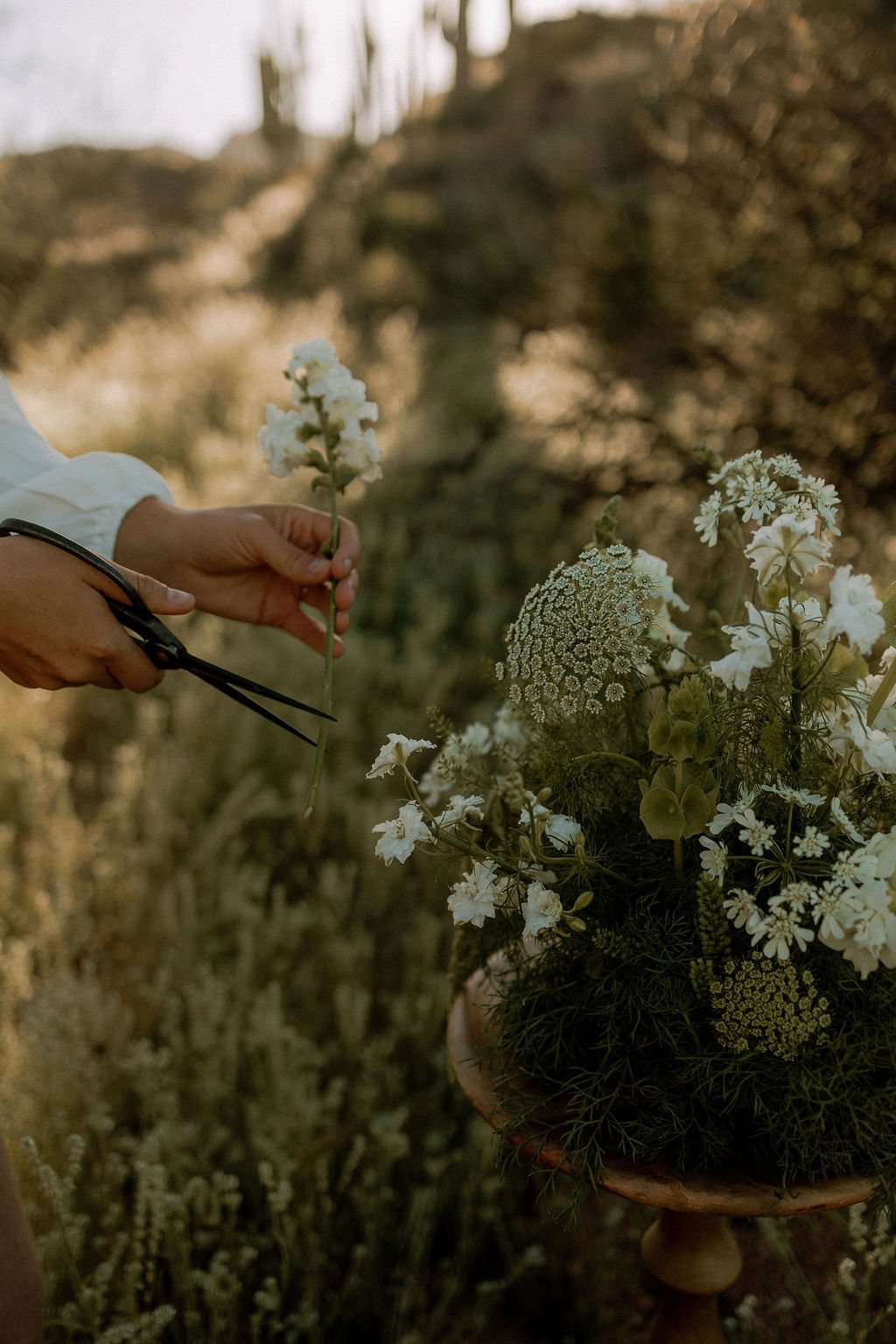 A person holding a white flower while preparing a bouquet of white and green flowers on a wooden pedestal outdoors.
