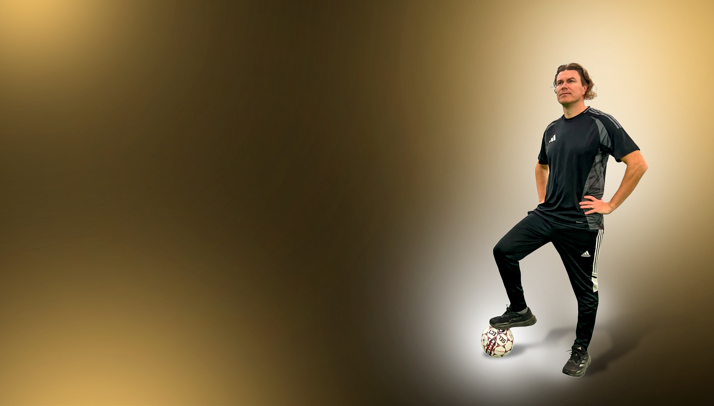 Mies seisoo jalkapallon päällä kädet lanteilla mustassa urheiluvaatteessa studion taustalla