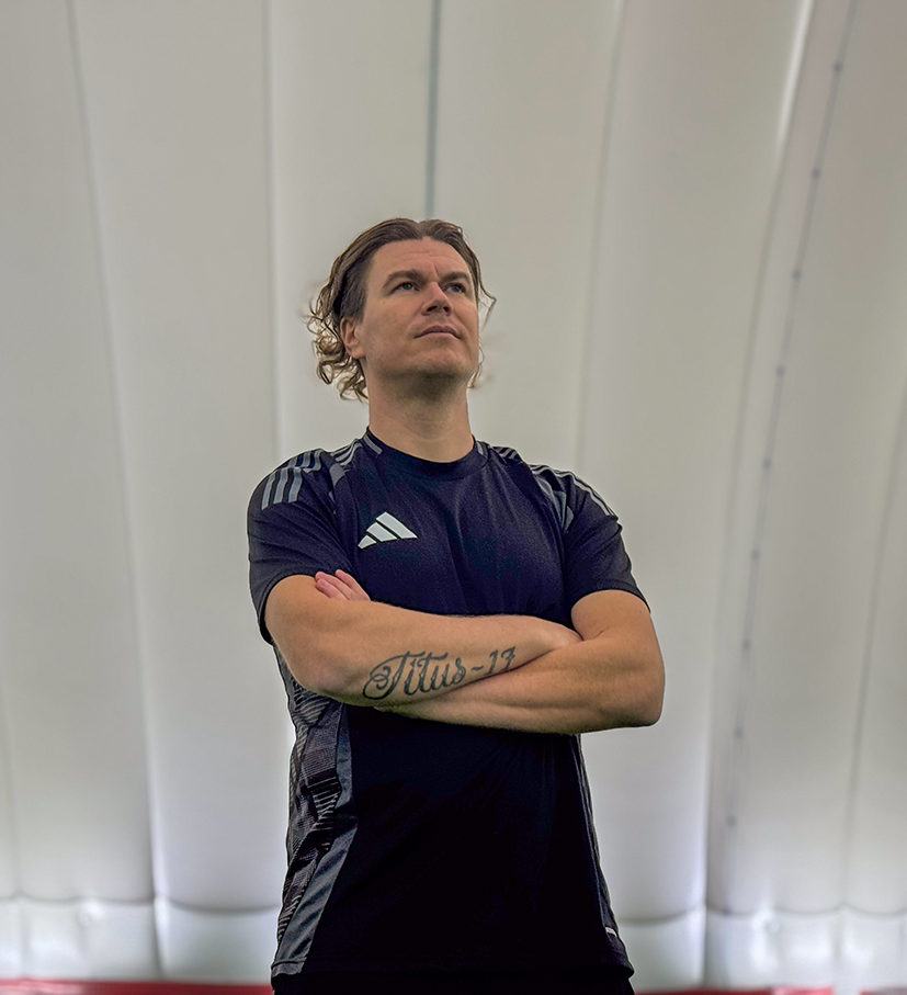 Nuori mies seisoo sisällä, katsoo ylöspäin ja kätensä ristissä rinnalla. Hänellä on tatuointi oikean kyynärvarren sisäpuolella, jossa lukee 'Titti—-17'.