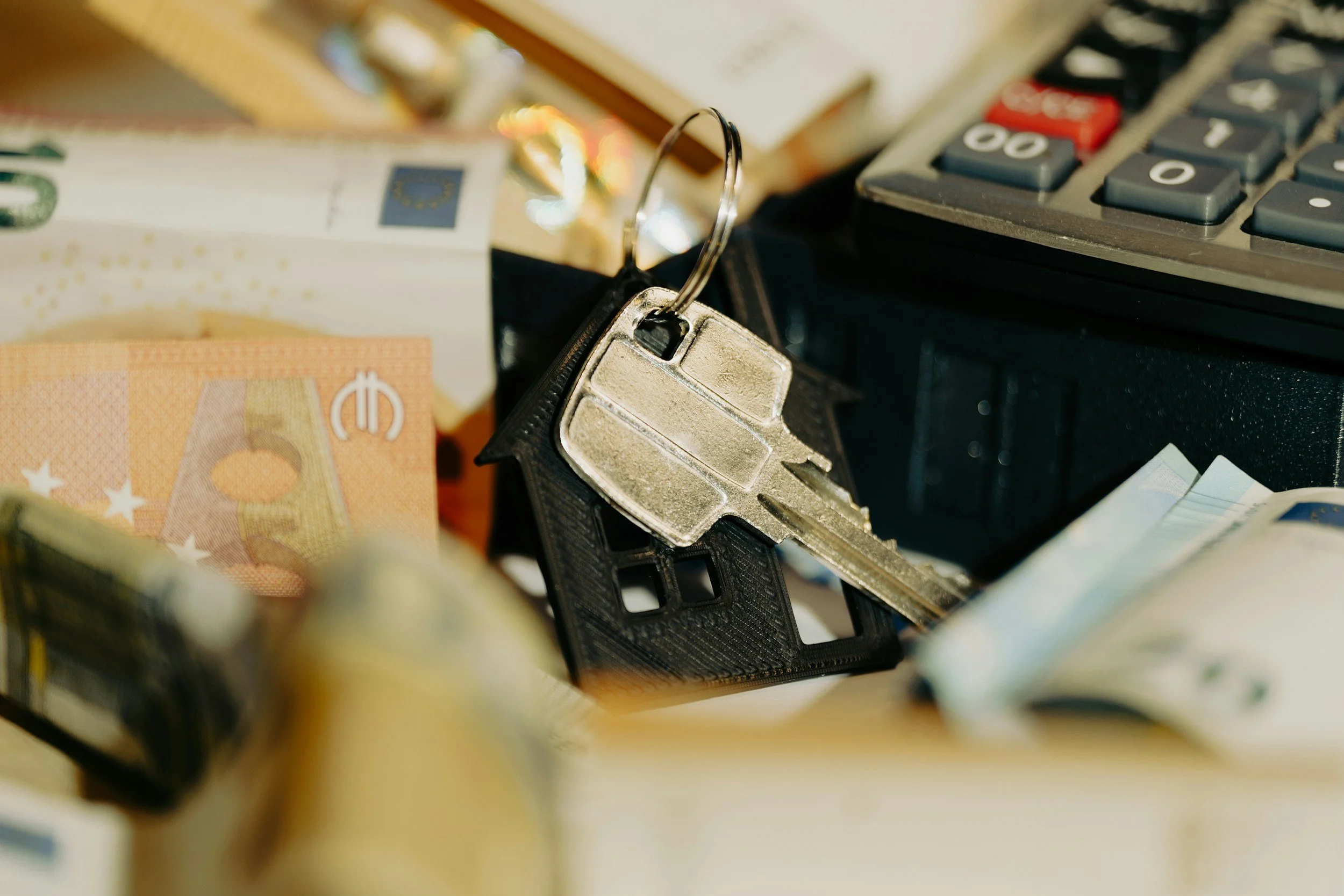 A keychain with a house-shaped key fob, placed on a desk with euro banknotes and a calculator.