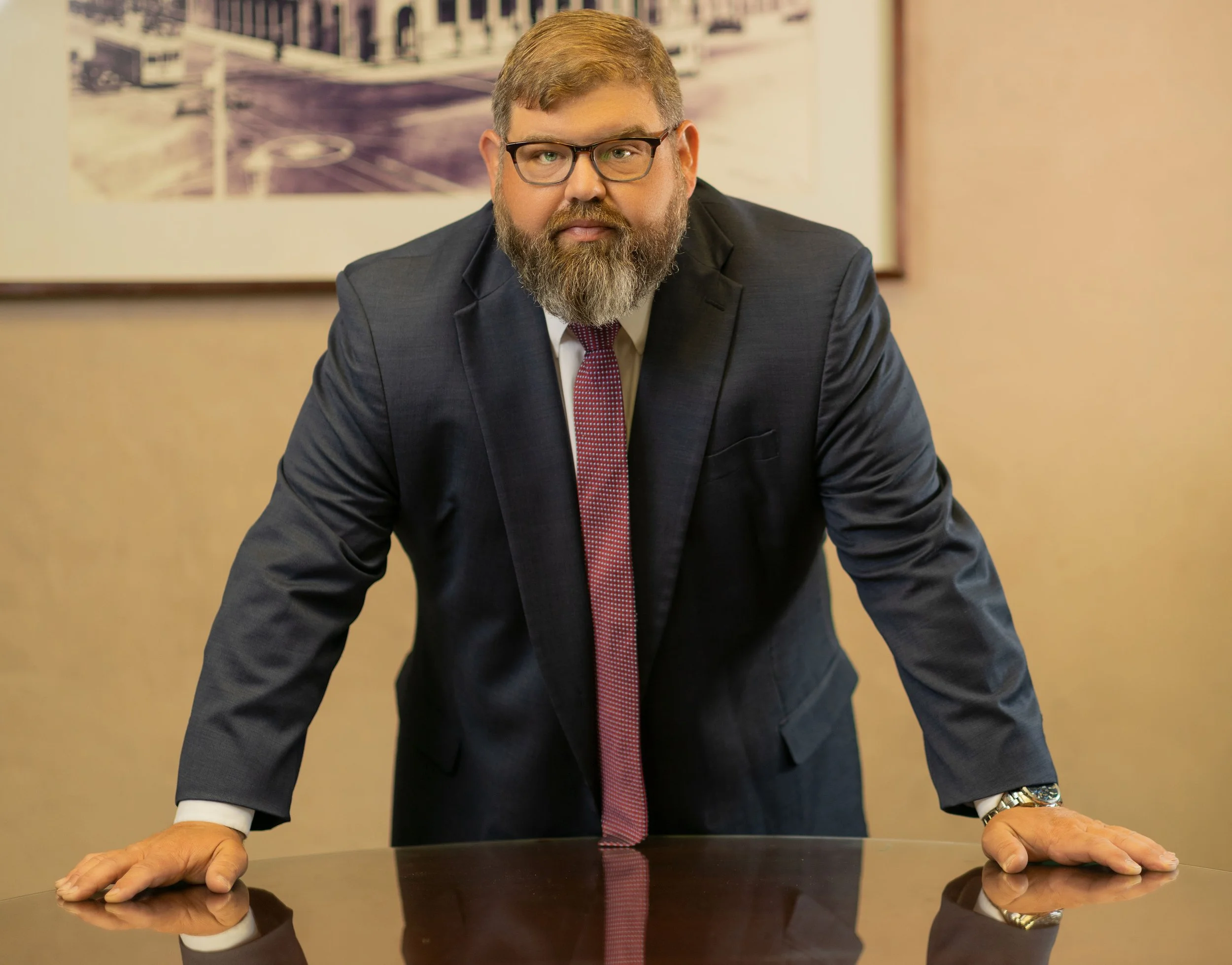 A man with glasses, a beard, and light brown hair, wearing a dark suit, white shirt, and red patterned tie, is standing behind a polished wooden table, leaning forward with his hands on the table. The background shows a framed black and white photograph of a basketball court.