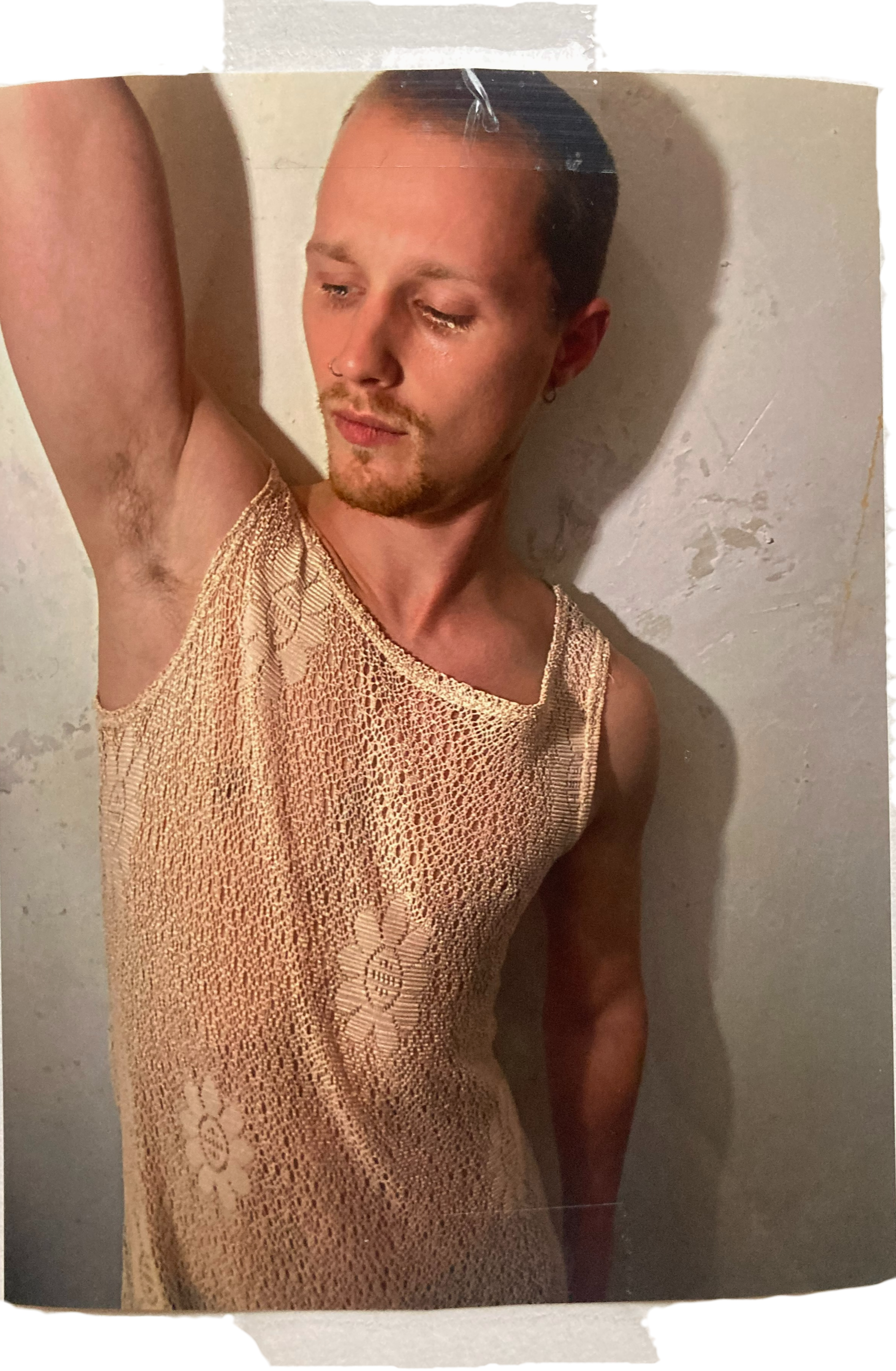 Individuell gestylter junger Mann mit Bart und Nasenpiercing trägt ein transparentes top mit Blumenmuster vor weißer Wand.