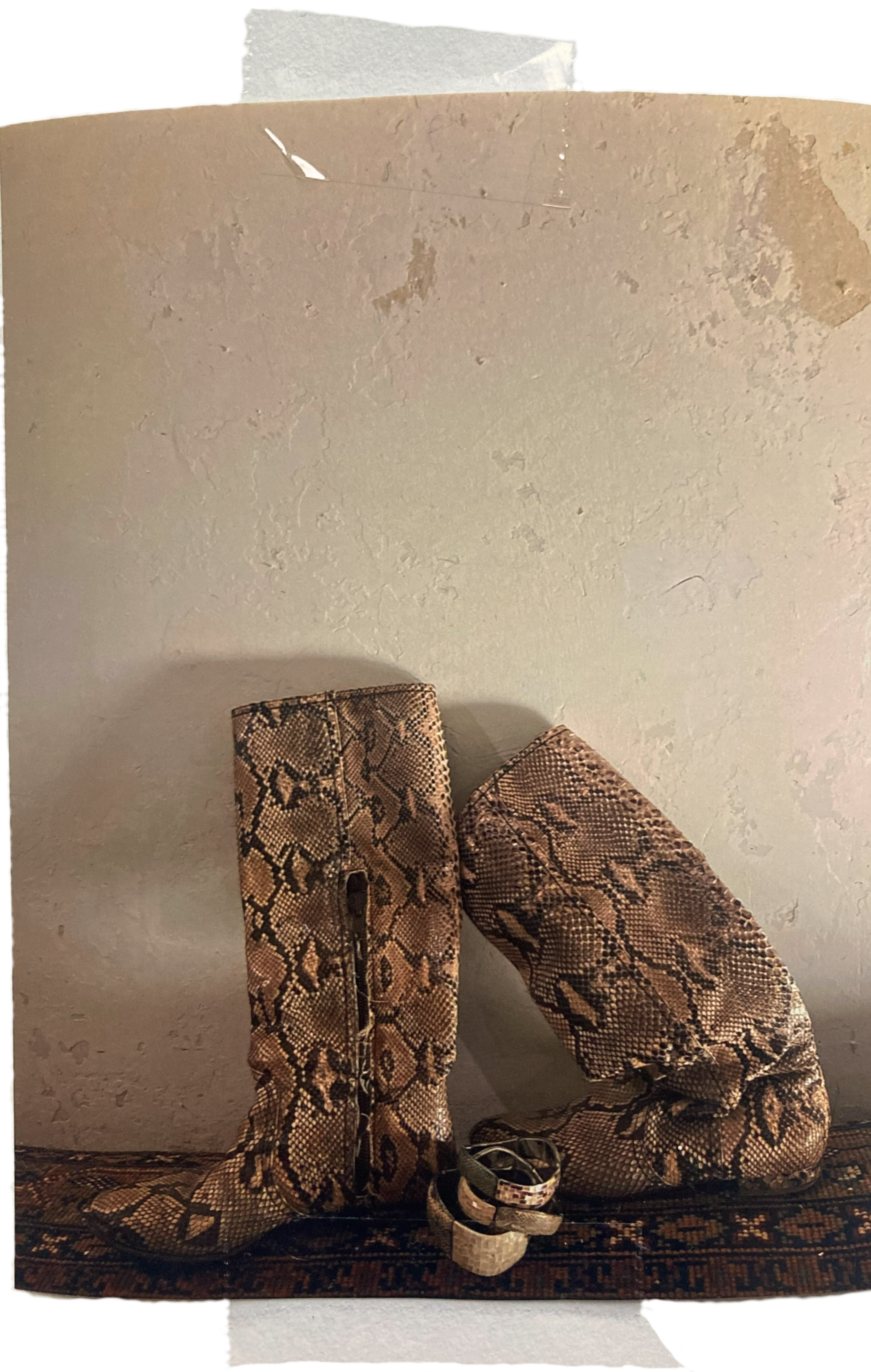Ein Paar Stiefel mit Schlangenledermuster und ein Gürtel auf einem Teppich, vor einer weißen Wand.