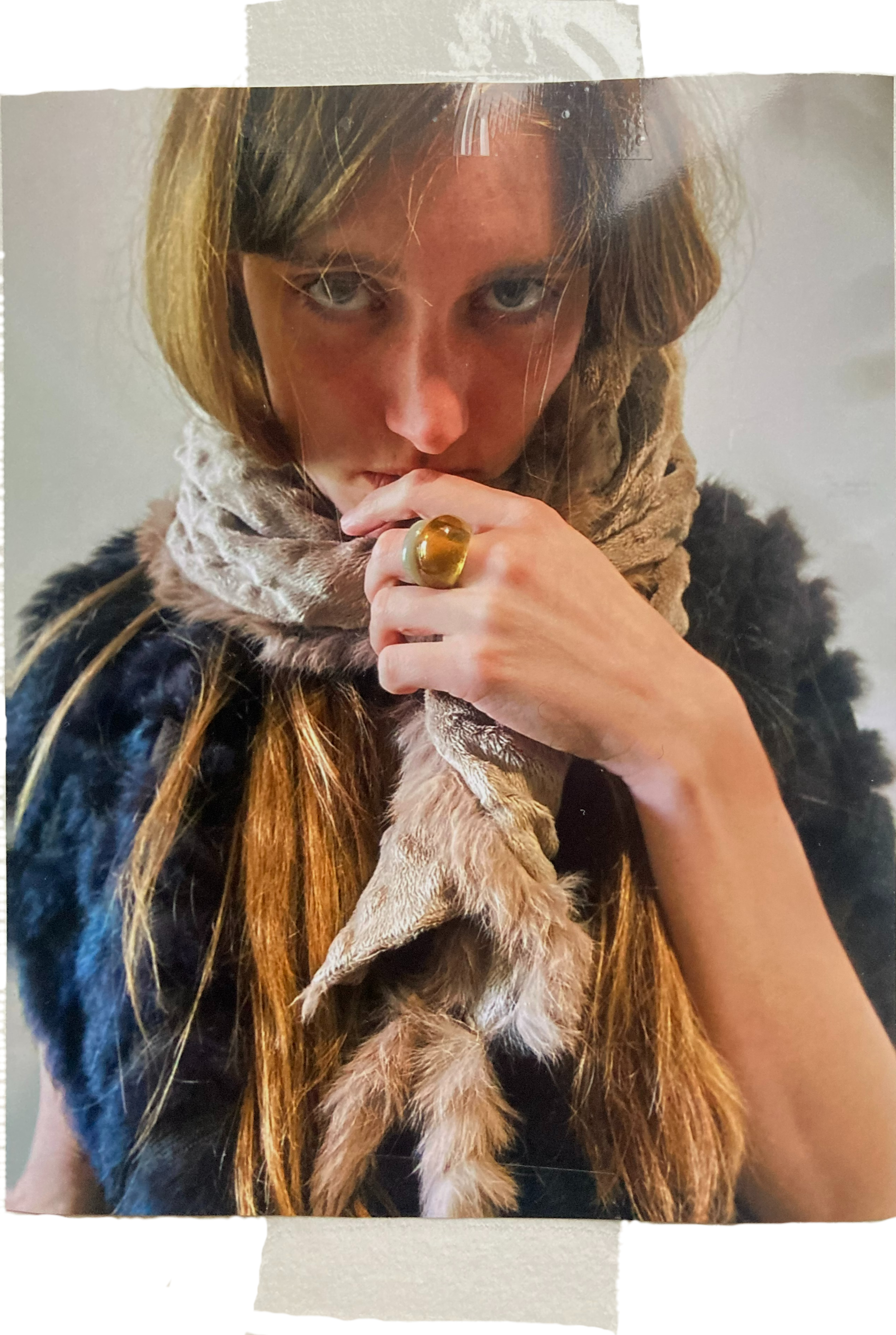 Junge Frau mit langen Haaren trägt einen Pelzmantel und einen Schal, hält ihre Hand mit einem großen goldenen Ring an den Lippen, schaut direkt in die Kamera.