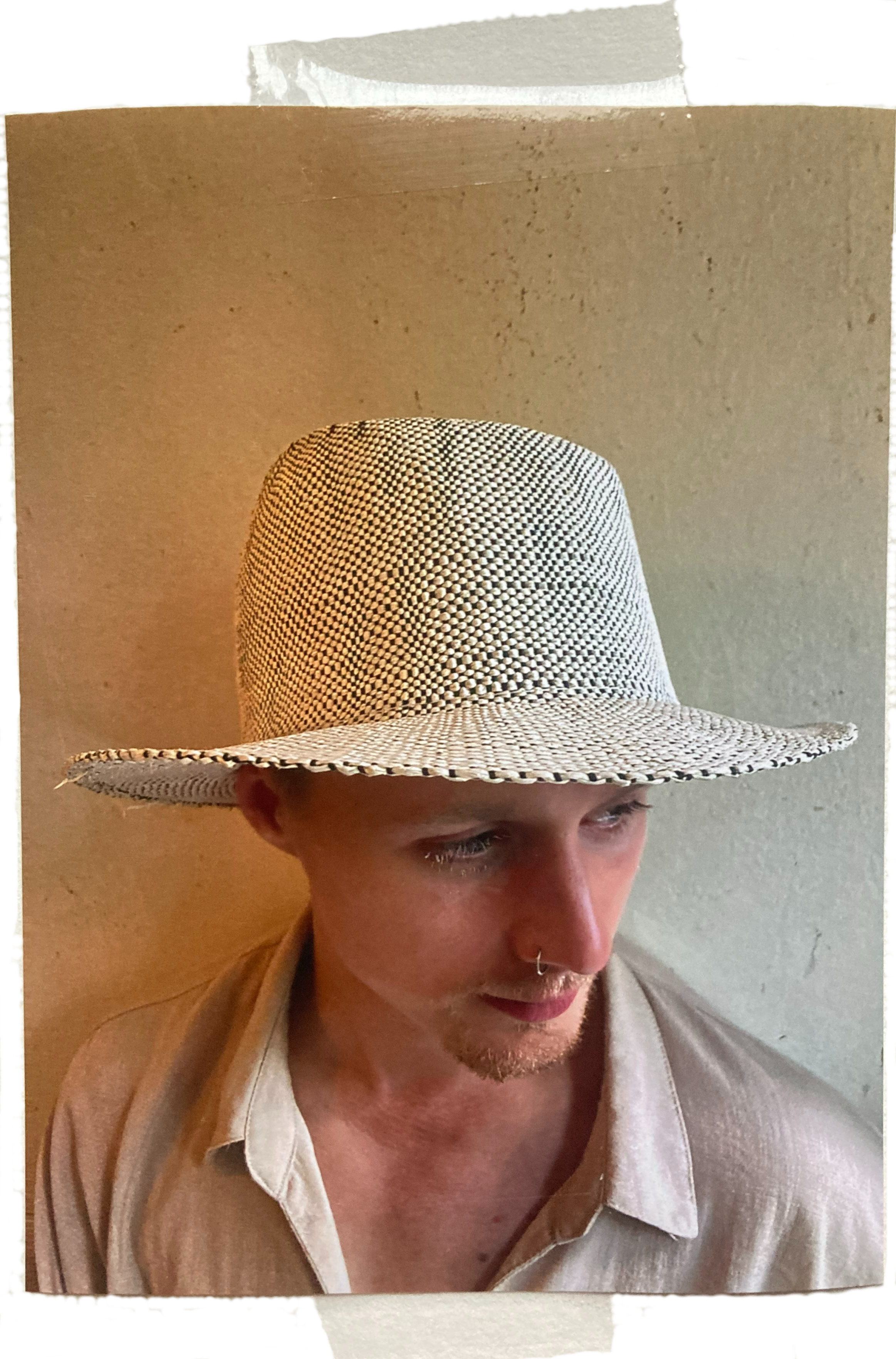 Individuell gestylter junger Mann mit Nasenpiercing trägt großen Strohhut vor neutraler Wand.