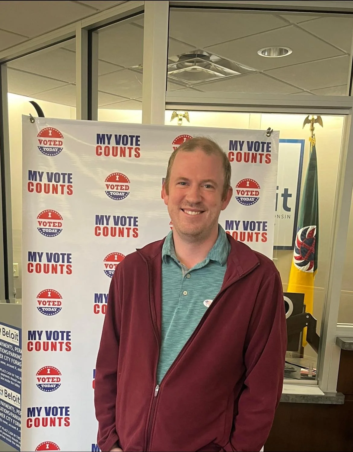 A man smiling at a voting location, standing in front of a backdrop with 'My Vote Counts' and 'Voted Today' logos, and an American flag visible behind him.