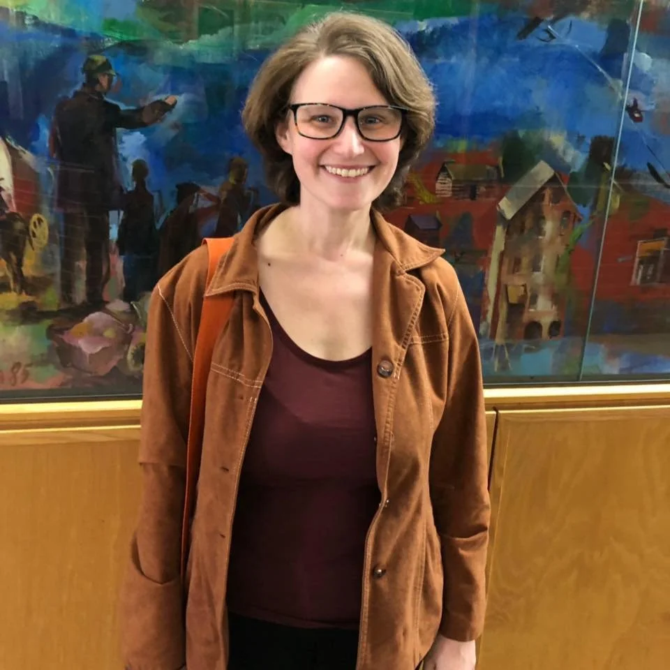 Woman with short brown hair and glasses smiling at camera, wearing a brown jacket and maroon shirt, standing in front of colorful abstract artwork.