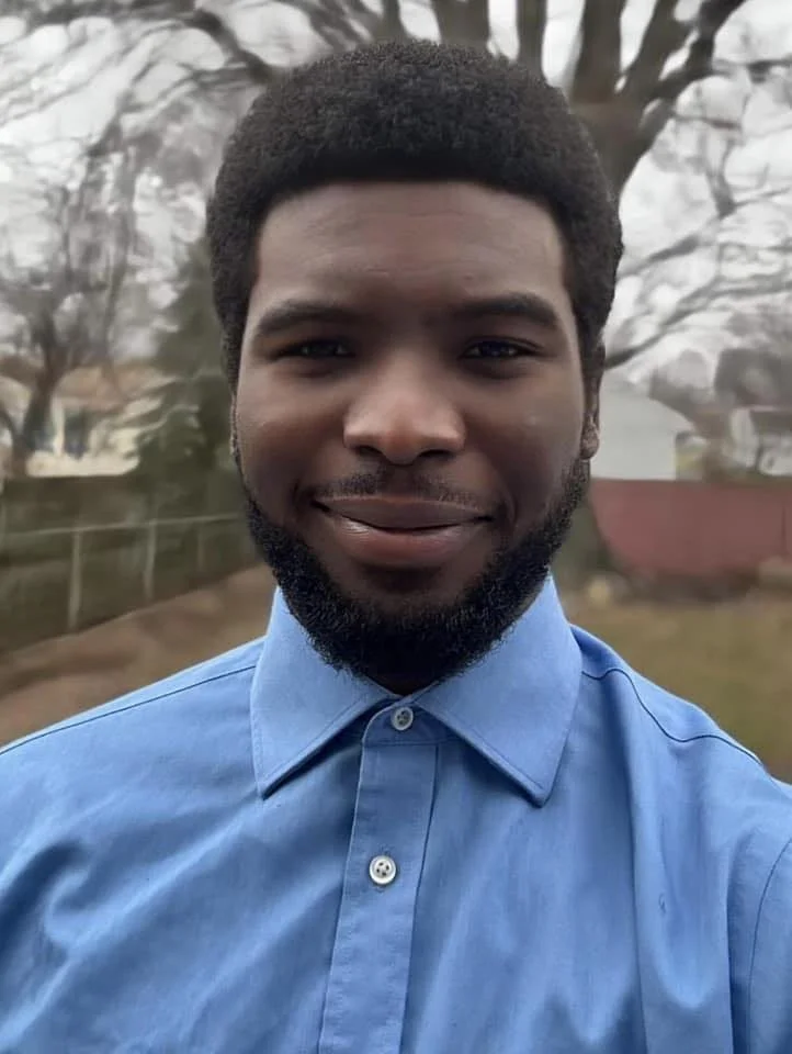 A young man with dark skin and a beard, wearing a blue button-up shirt, smiling outdoors with trees and a fence in the background.