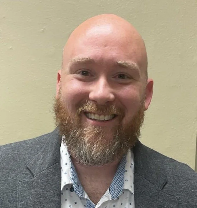 Smiling bald man with a beard wearing a suit jacket and a white shirt with small dots, standing against a plain wall.