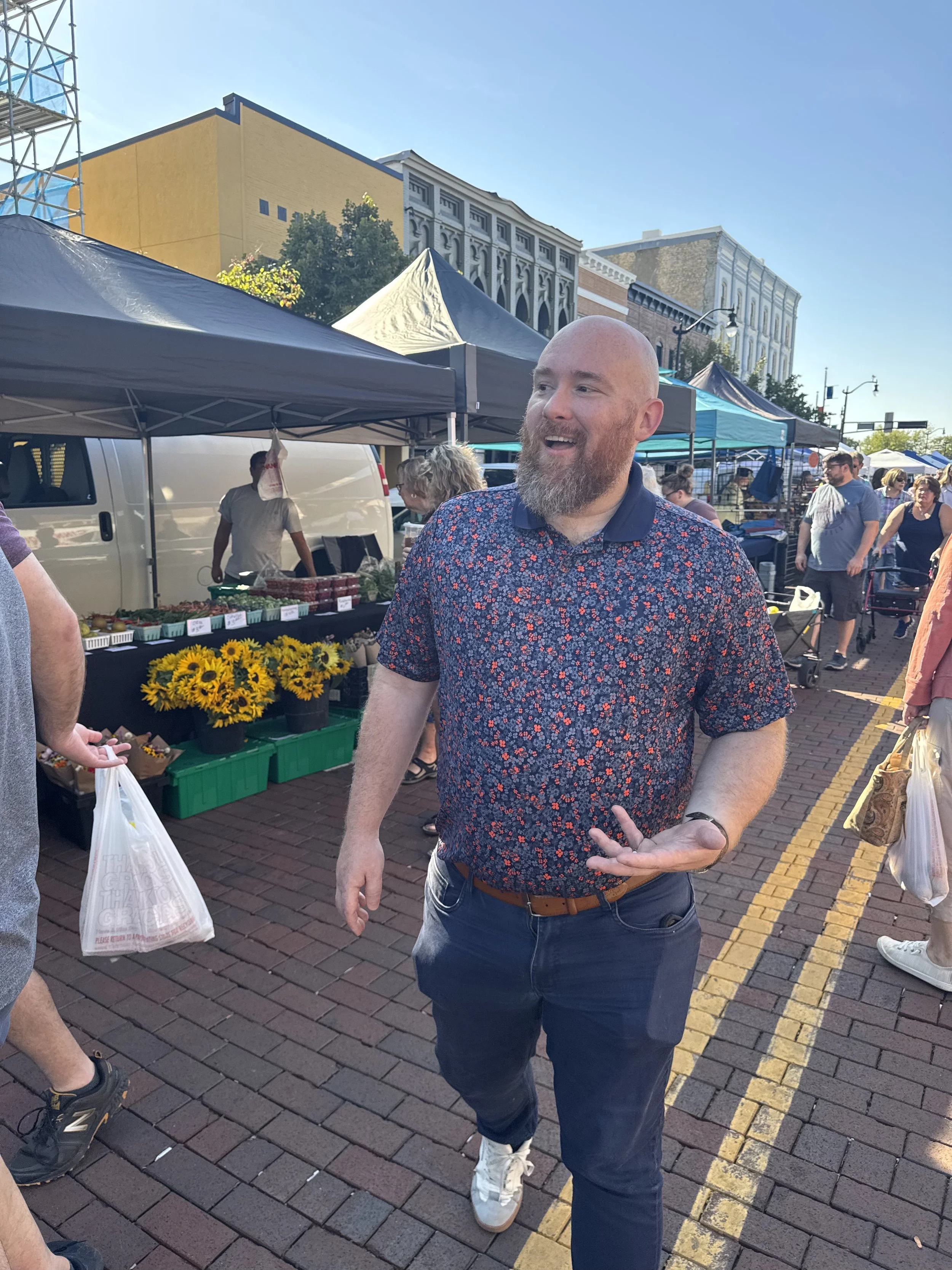 A man with a beard wearing a floral shirt and dark jeans standing and smiling at a market with tents and other shoppers in the background.