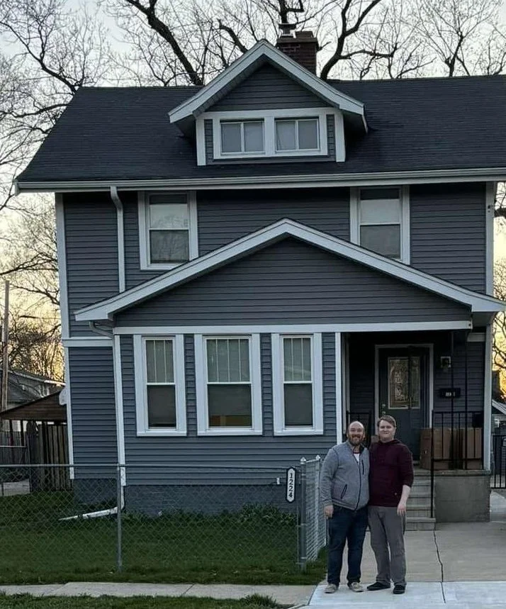 A two-story blue house with white trim and a front porch, two people standing on the sidewalk in front of it.